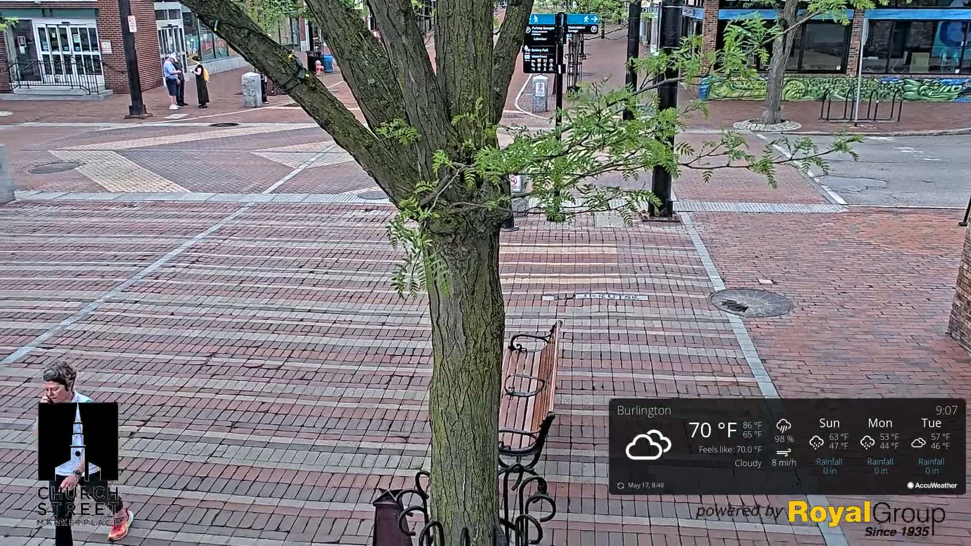 A brick paved city street scene under cloudy skies shows a tree, a park bench, and several pedestrians.