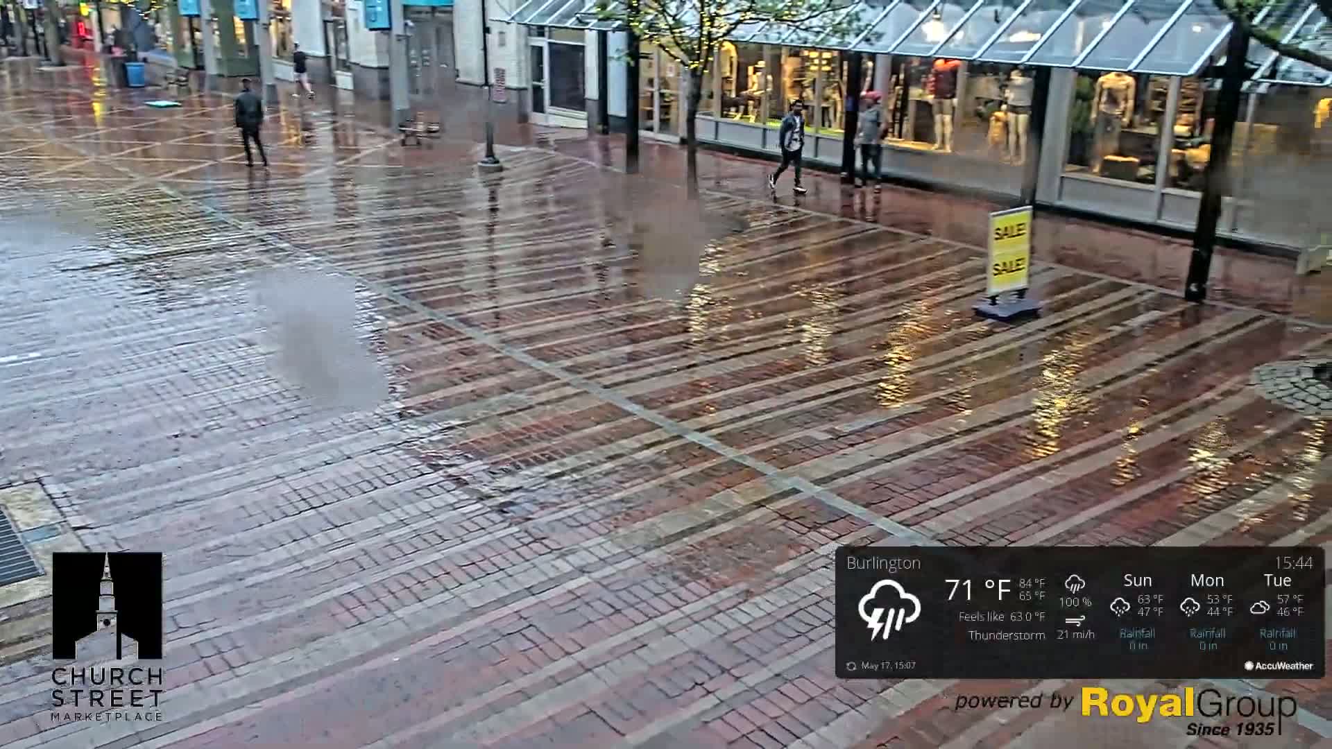 A wet brick-paved street, lined with storefronts, is sparsely populated with pedestrians during a thunderstorm.
