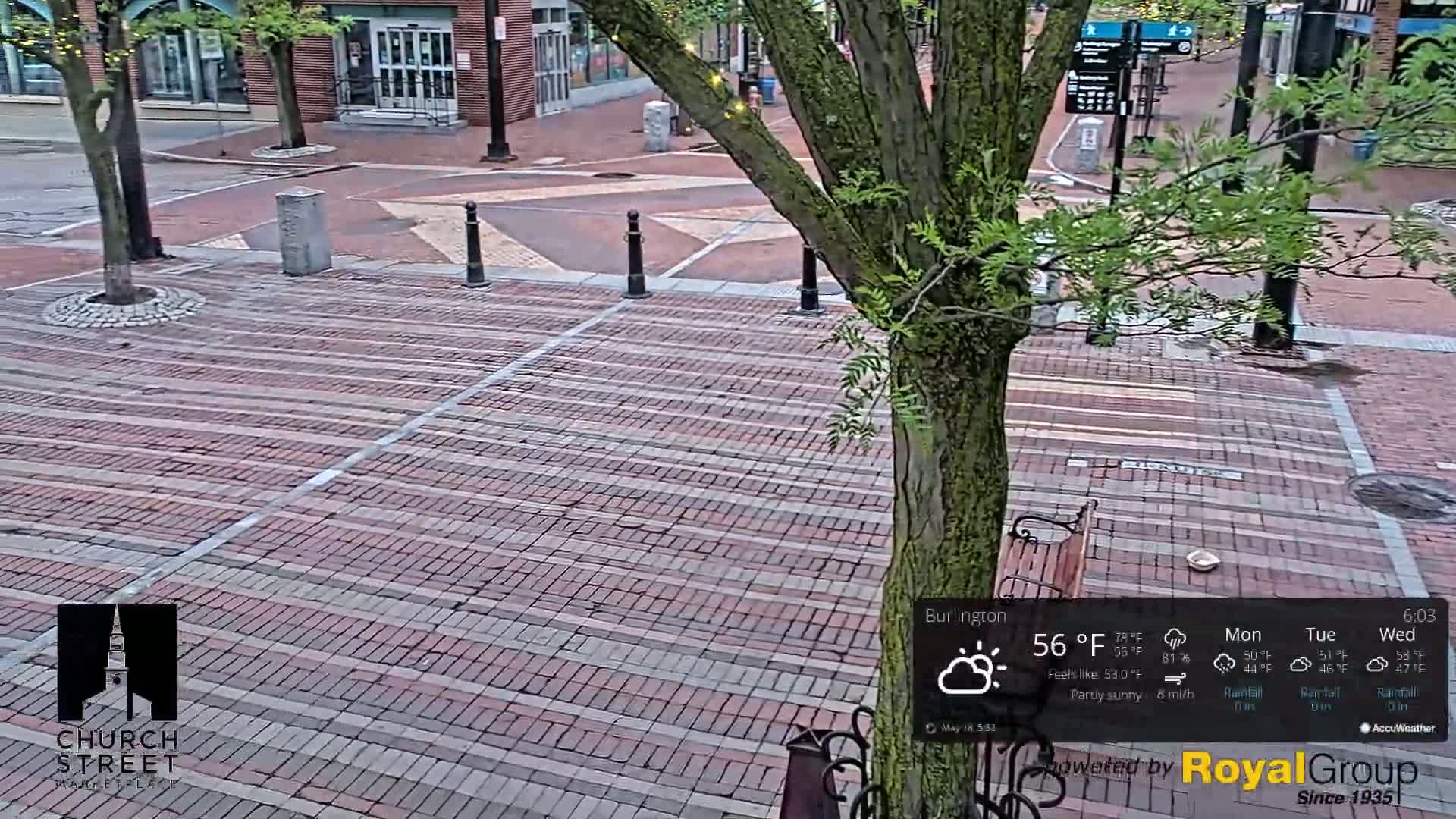 A brick paved plaza with a tree in the center, partially sunny weather conditions.