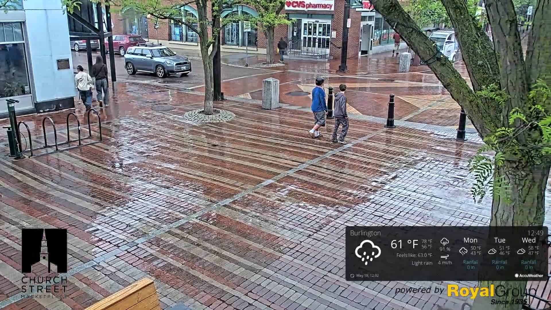 A brick pedestrian area is wet from light rain, and several people are walking among the trees and buildings.