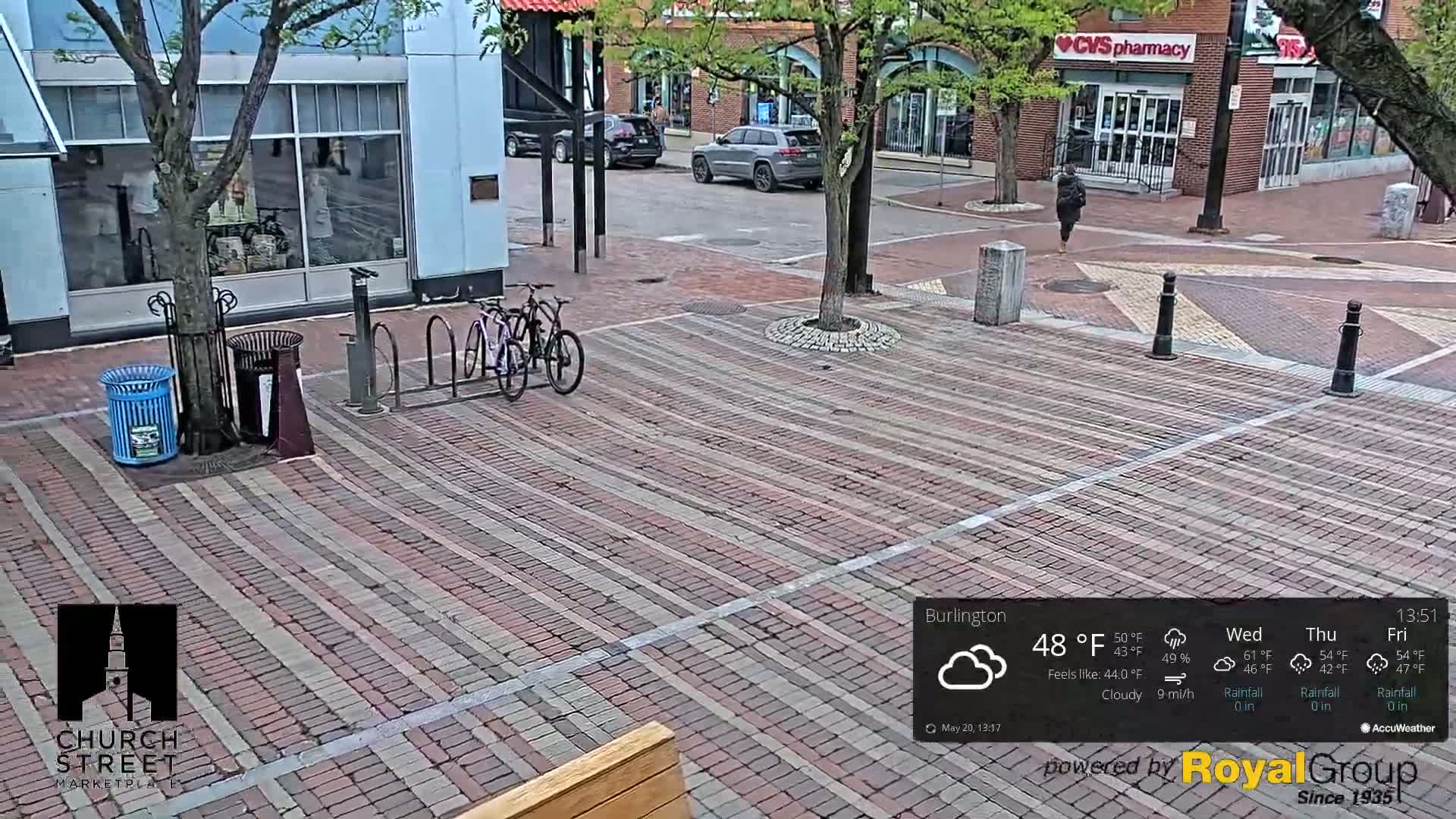 A brick paved street scene on a cloudy day shows two bicycles at a bike rack, a person walking in the distance, and some parked cars.