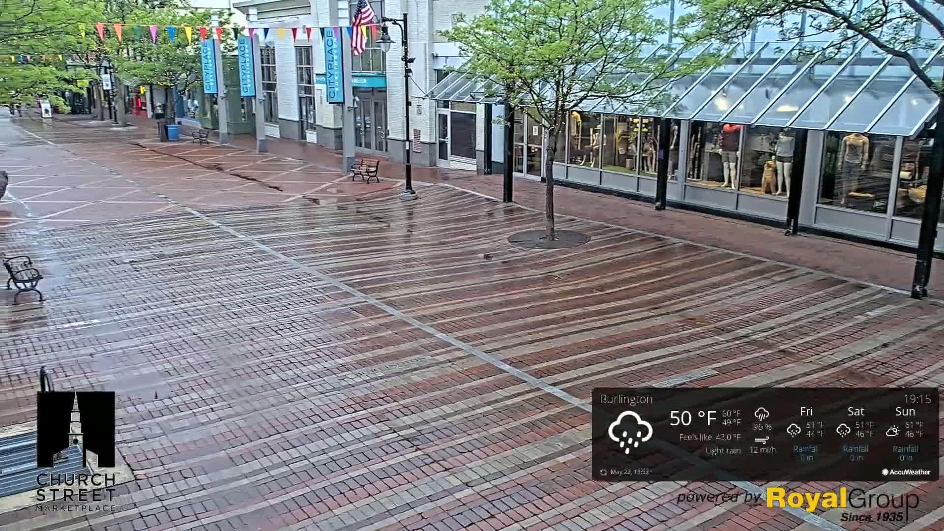 A wet brick pedestrian walkway stretches between shops under a light rain.