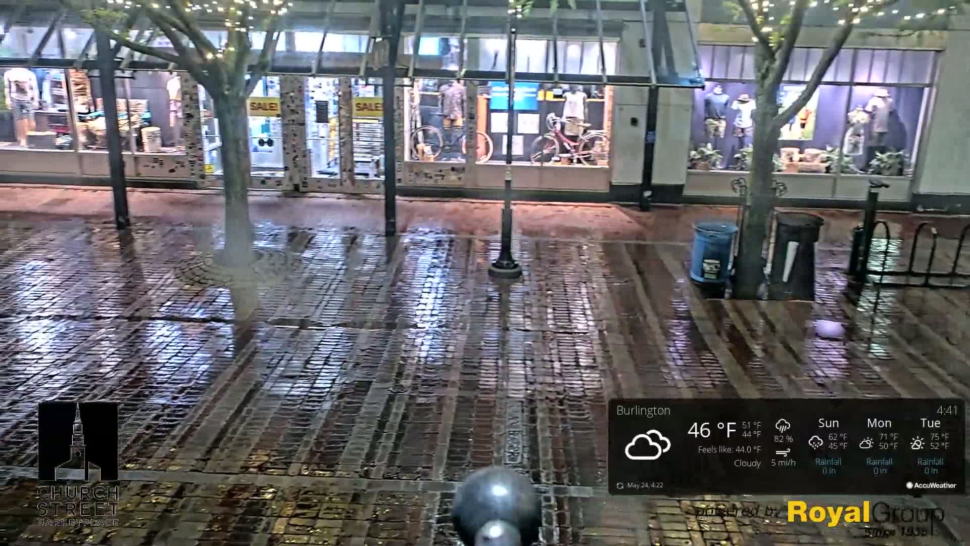 A wet brick street at night is seen in front of several storefronts with mannequins displayed in the windows under a cloudy sky.