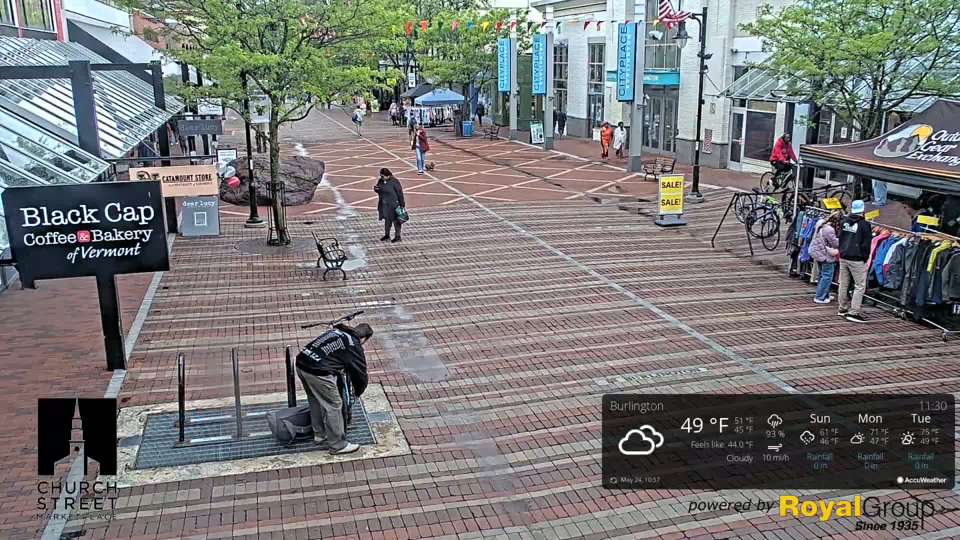 A brick pedestrian walkway in a city, under cloudy skies with light rain, features several people walking and shopping near outdoor vendors and bike racks.