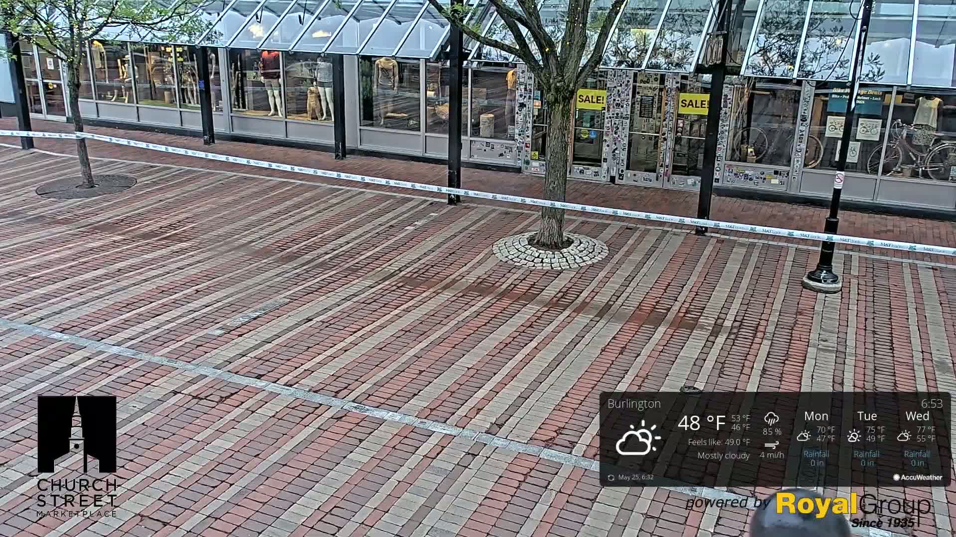 A mostly cloudy day shows a brick paved street in front of shops with a tree in the center, a light rain is falling.
