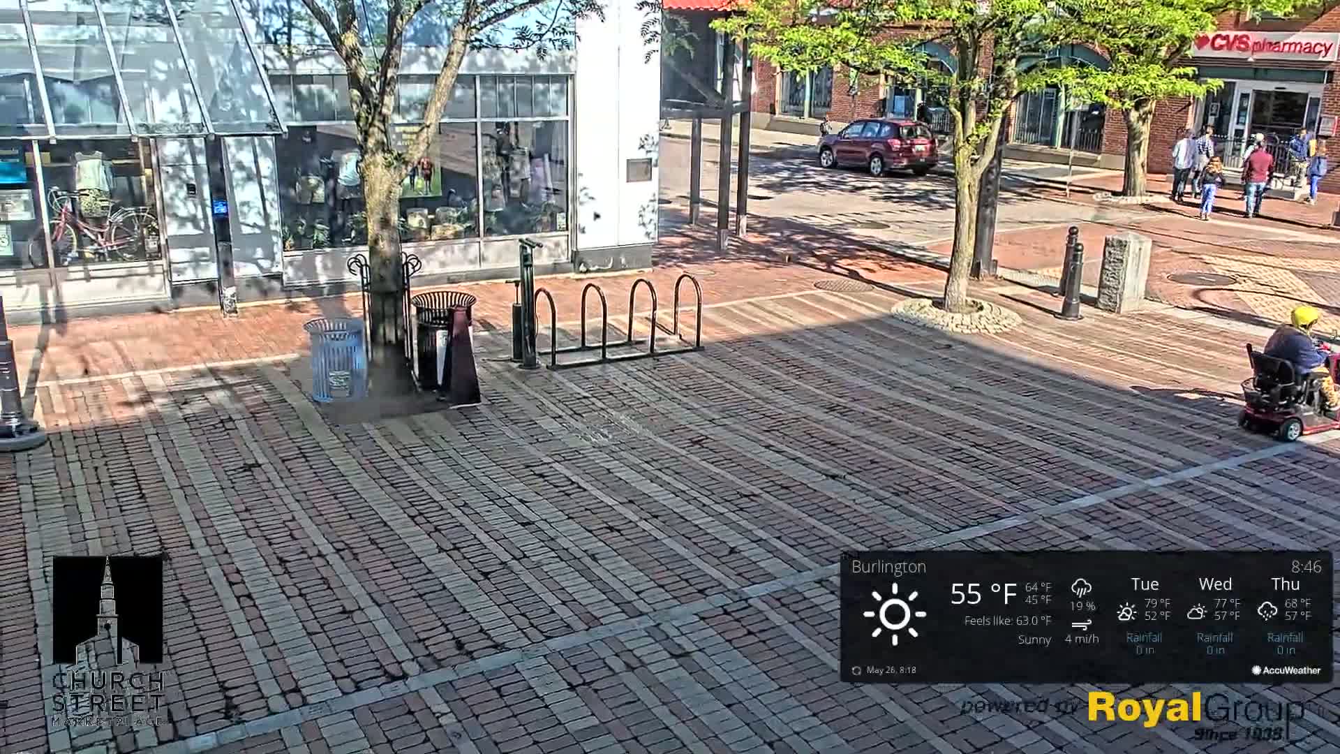 A brick-paved city square on a sunny day, with a person in a mobility scooter, a bike rack, and trash cans.