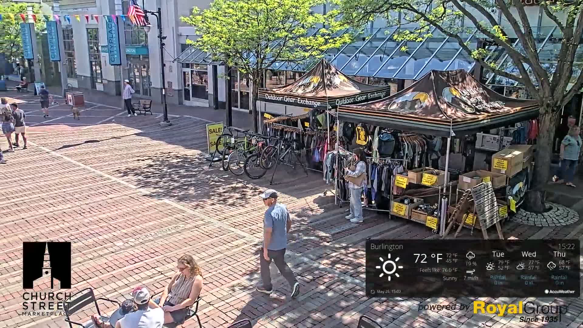 A brick marketplace with a pop-up shop selling outdoor gear, several pedestrians, and sunny weather.