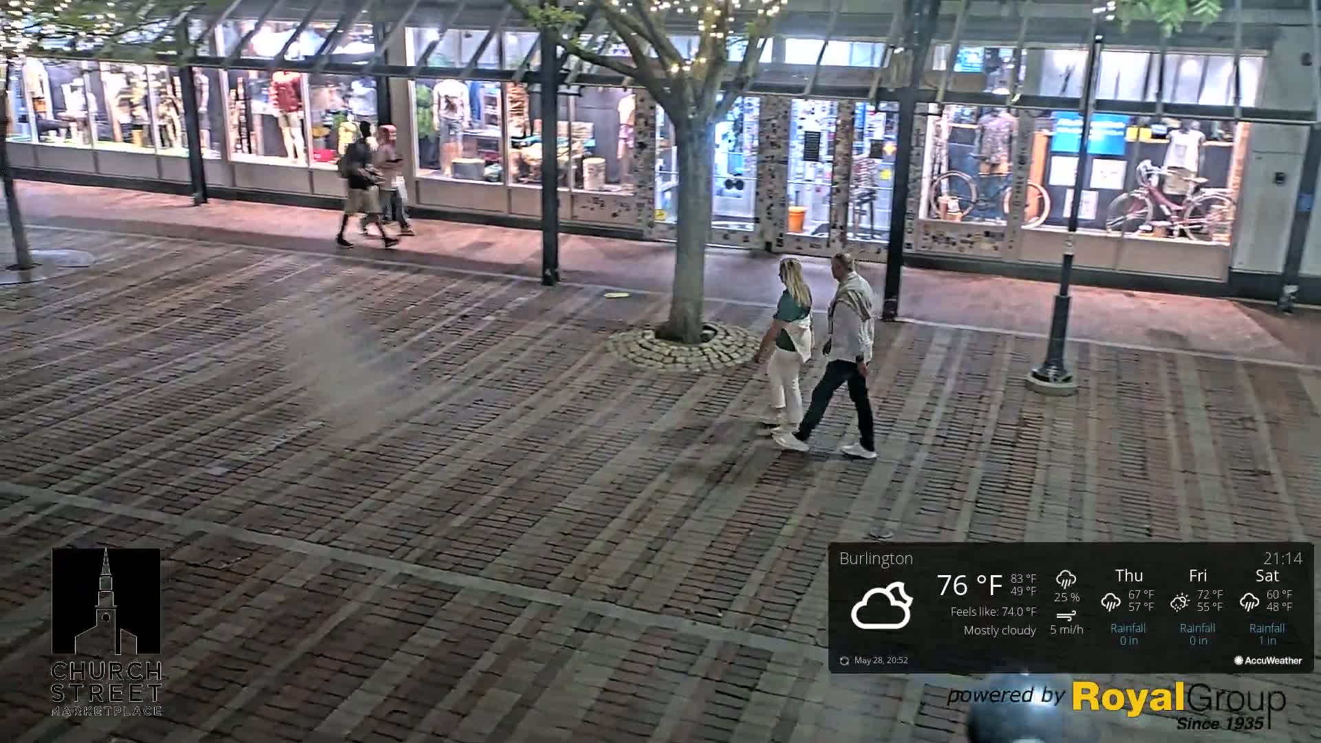 A brick paved area at night under mostly cloudy skies, with a few people walking past illuminated storefronts.