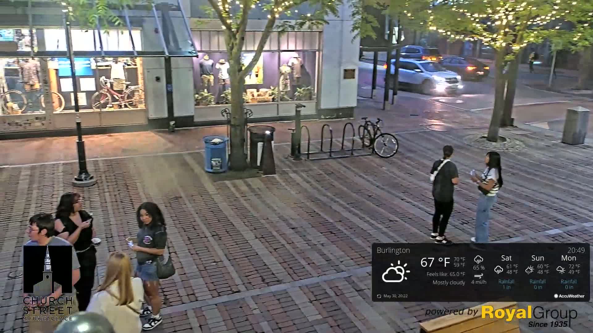A mostly cloudy evening scene shows several people standing on a brick paved street in front of shops, with a bicycle rack and a parked bicycle visible.