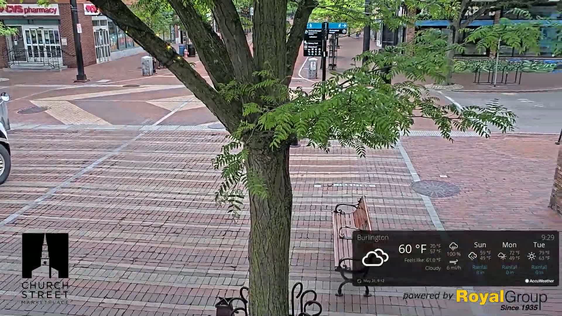 A brick-paved street scene with a tree in the foreground, a park bench, and cloudy weather at 60°F.