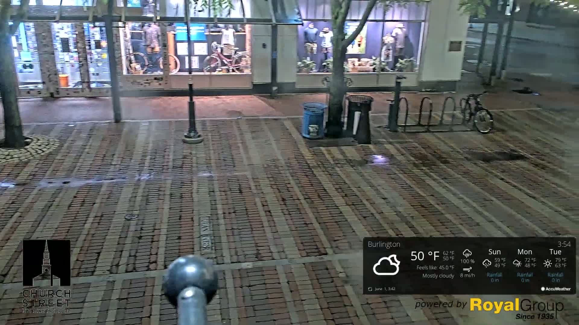 A wet brick street at night, under mostly cloudy skies, with storefront displays visible and a bicycle parked nearby.