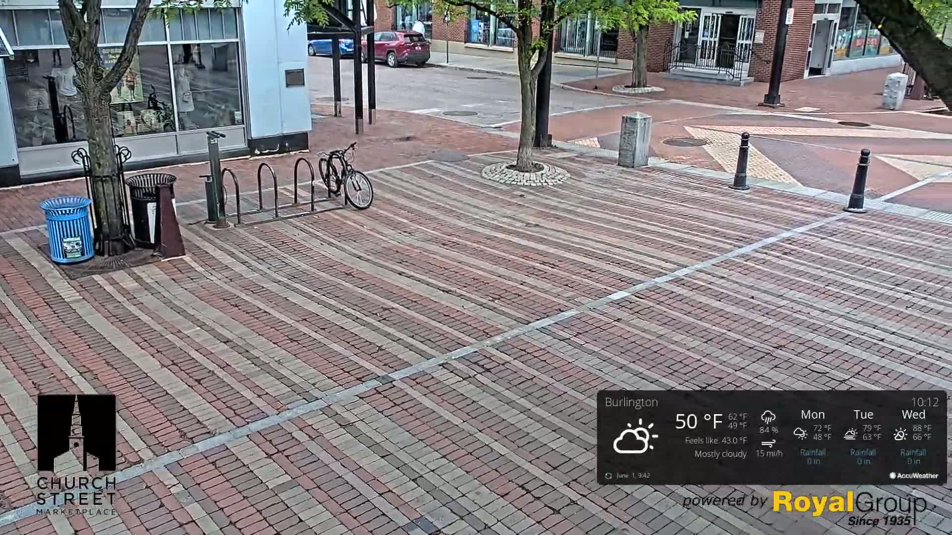 A mostly cloudy, brick-paved street scene shows a bicycle locked to a rack near a tree, trash cans, and a few parked cars in the distance.