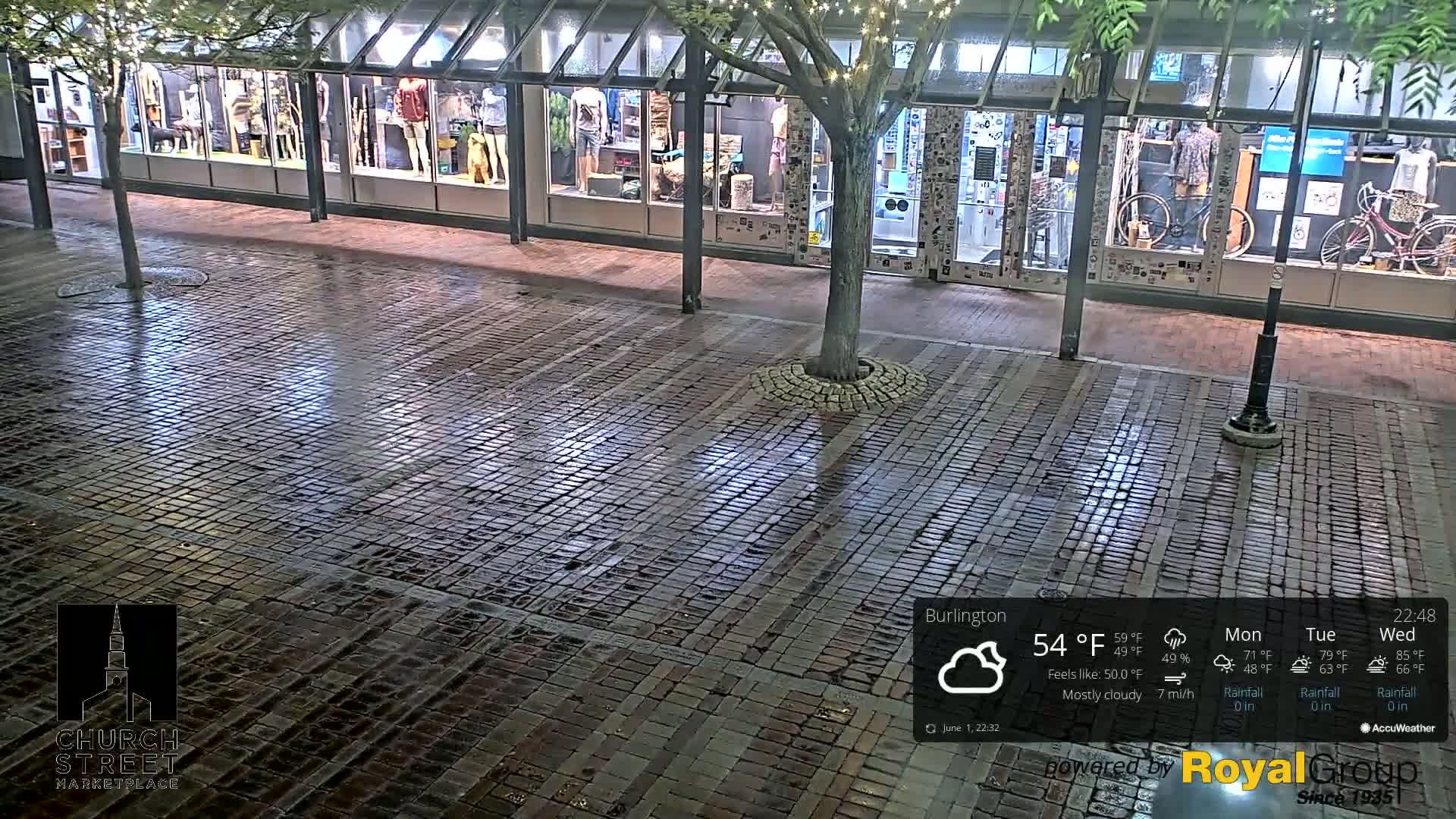 A wet brick pavement at night, under mostly cloudy skies, in front of a storefront with several display windows.