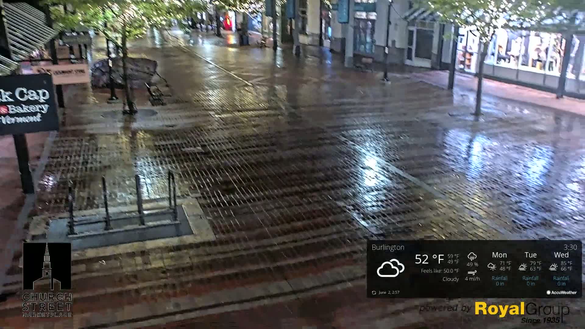 A wet brick-paved street is seen under cloudy conditions at night, with reflections of lights visible on the slick surface.