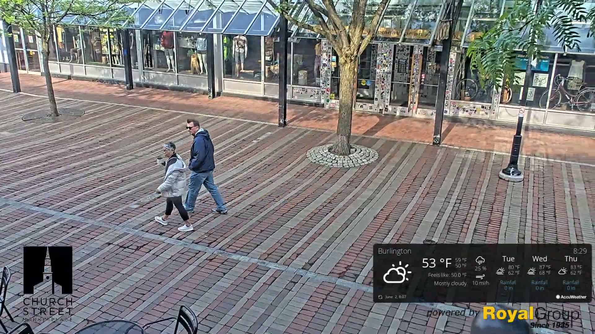A man and woman walk across a brick paved street under mostly cloudy skies.