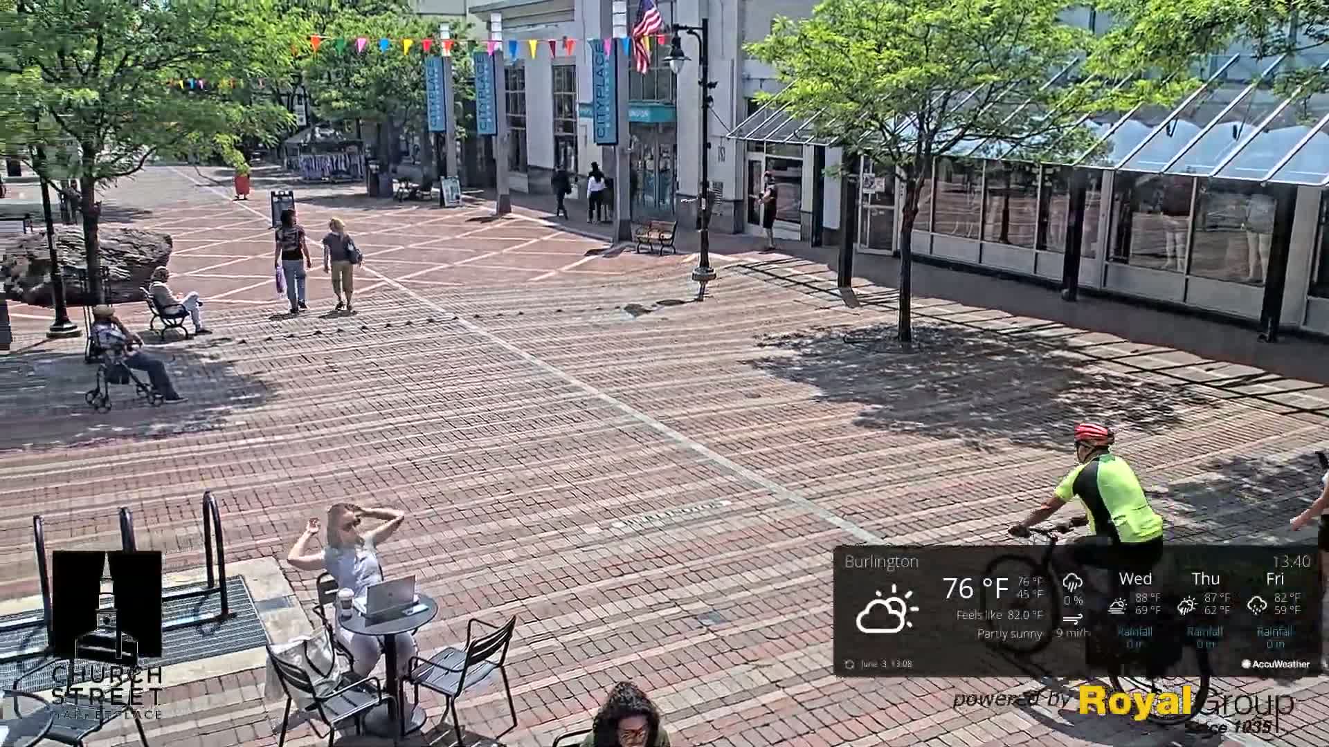 A brick plaza with several people walking and sitting, some on benches and some at outdoor tables, under partly sunny conditions.