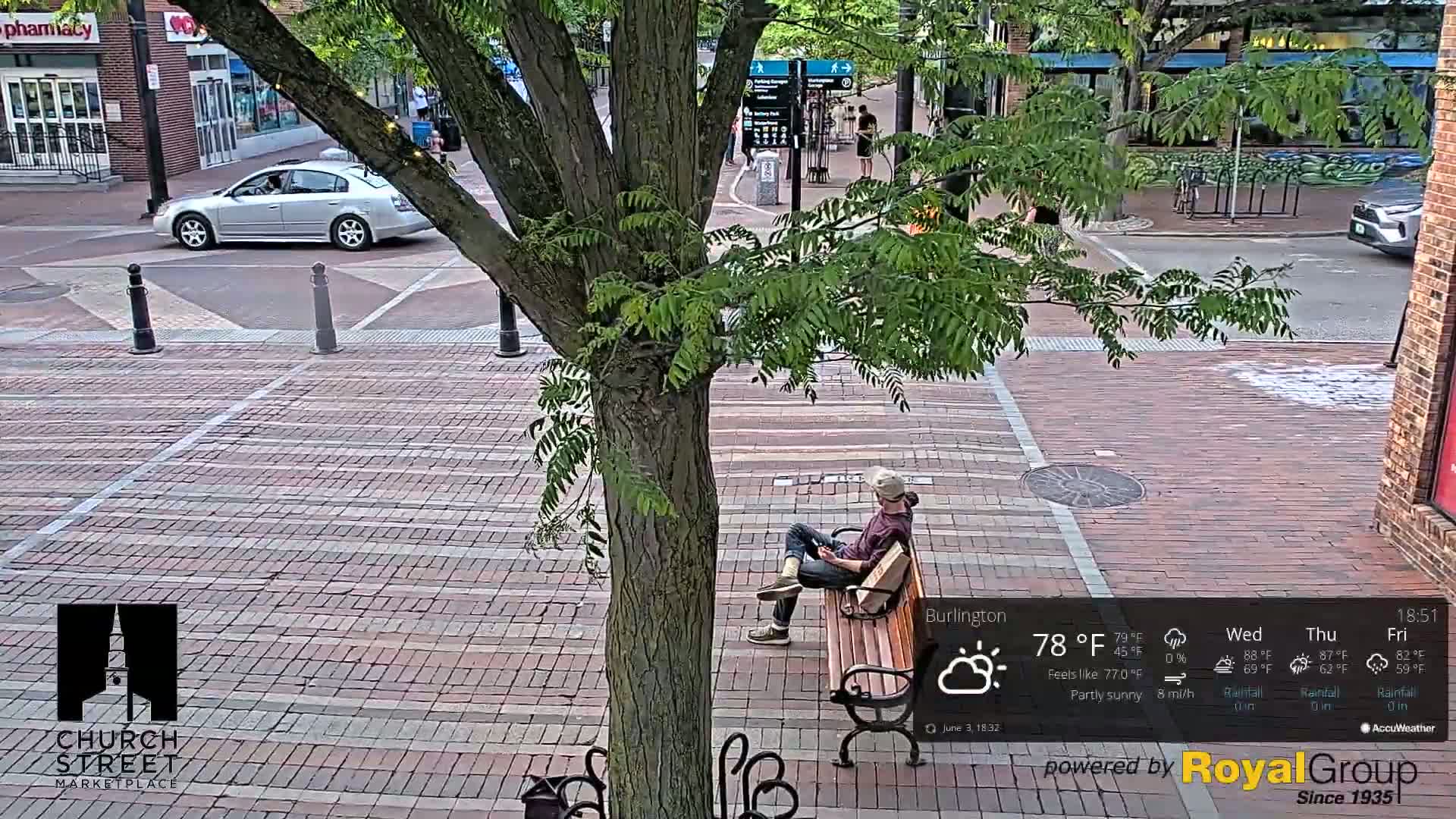 A person sits on a park bench on a partly sunny brick street, with a silver car parked nearby.