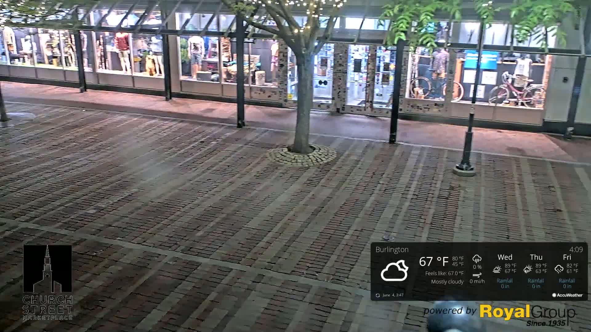 A brick-paved plaza at night, mostly cloudy, with a tree in the center and store fronts visible in the background.