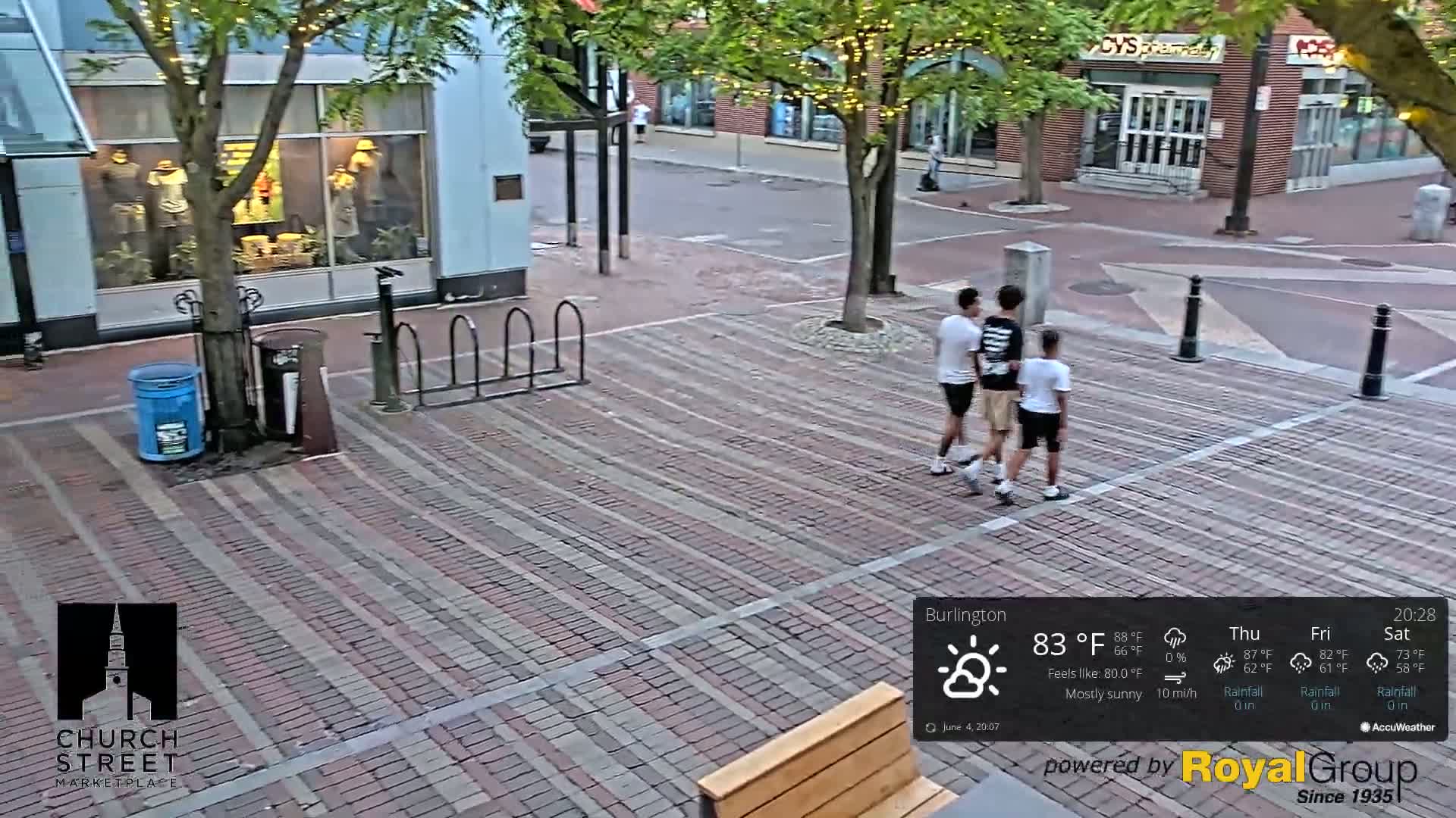 Three people walk on a brick-paved street under a mostly sunny sky.