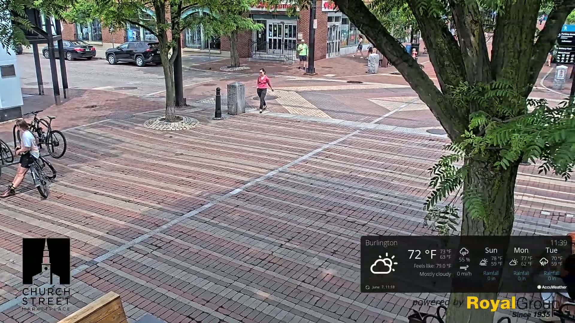 A mostly cloudy day shows a brick-paved city square with a person on a bicycle, another person walking, and several parked cars visible in the background.