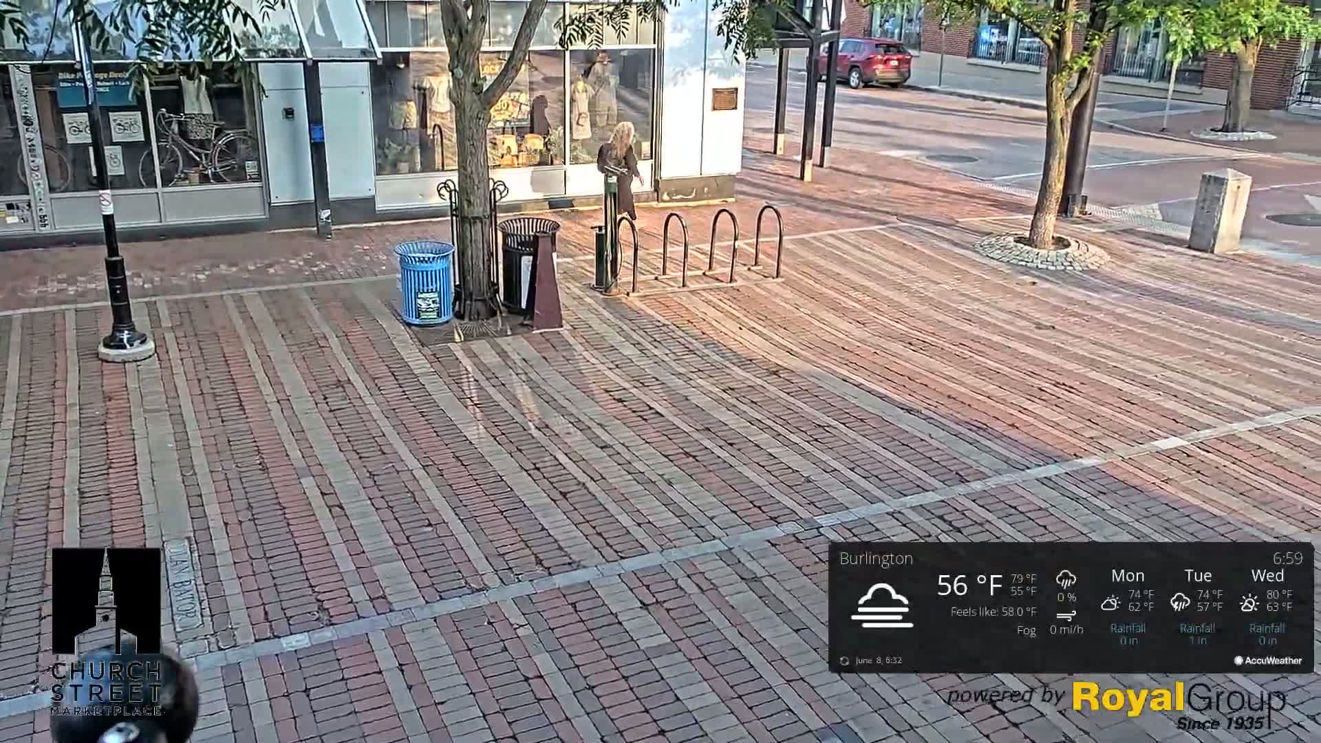 A brick paved area, partially shaded by a tree, shows a person standing near a bike rack and trash cans on a partly cloudy day.