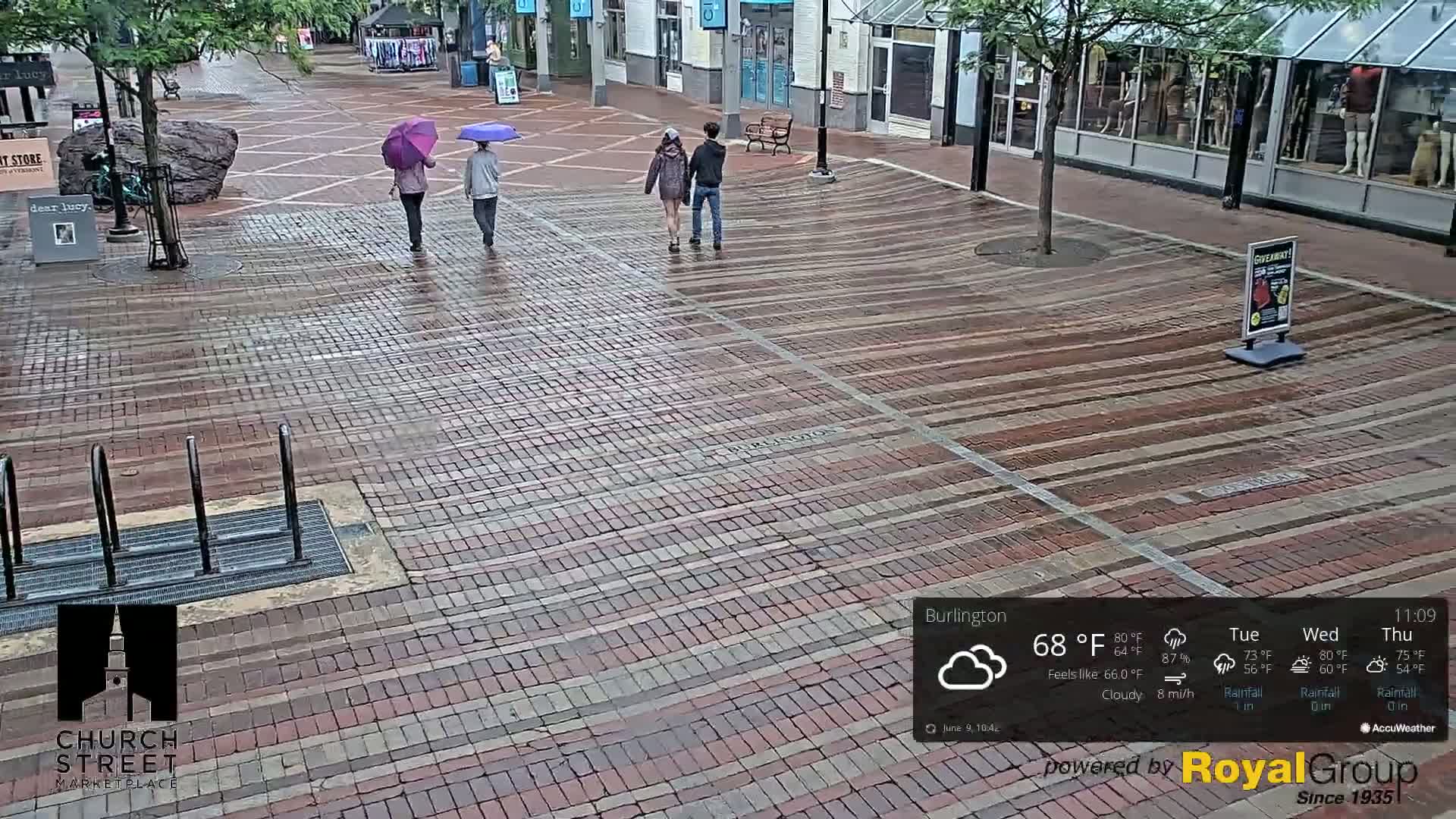 Three people with umbrellas walk on a rain-slicked brick street.