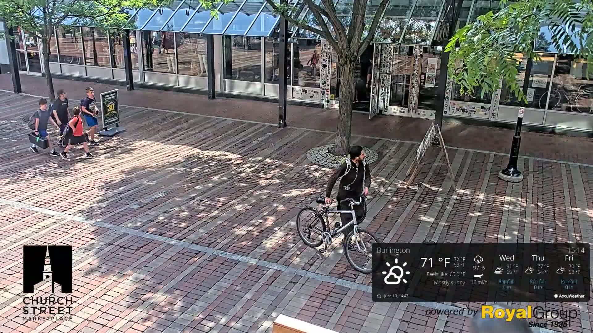 A mostly sunny outdoor scene shows a brick paved area with pedestrians and a person riding a bicycle; the shadows indicate the time of day.