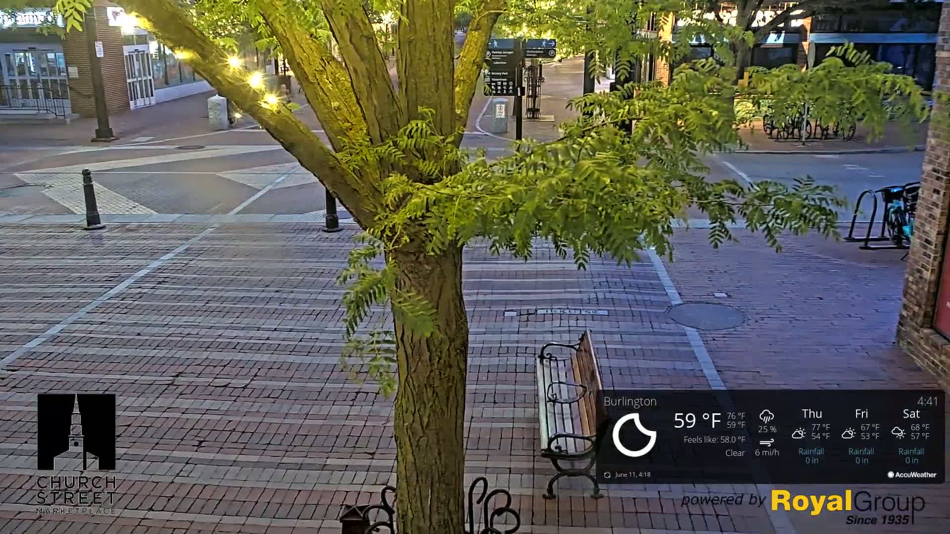 A brick-paved street scene at dusk, under a clear sky, features a tree with string lights, a park bench, and bicycle racks.