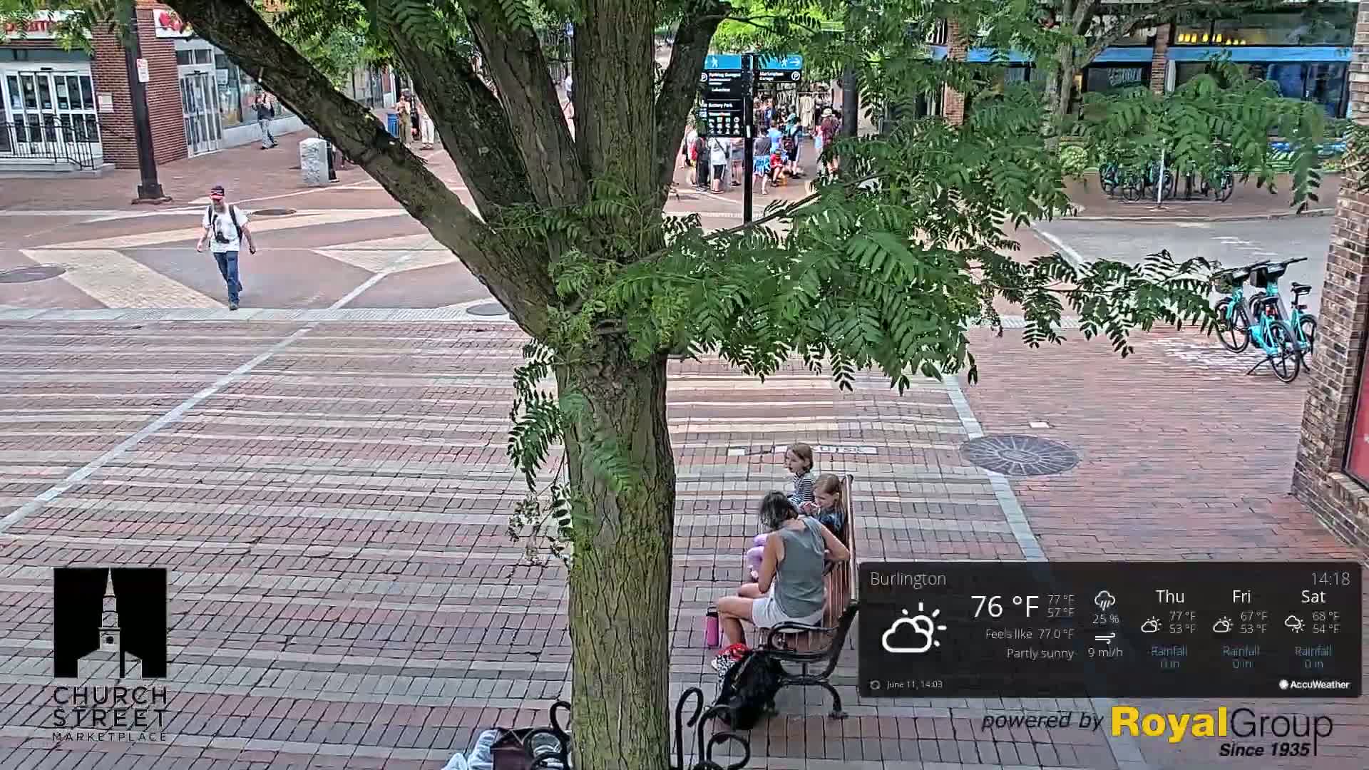 A partly sunny outdoor scene shows a brick plaza with a tree, several pedestrians, a family sitting on a bench, and parked bicycles.