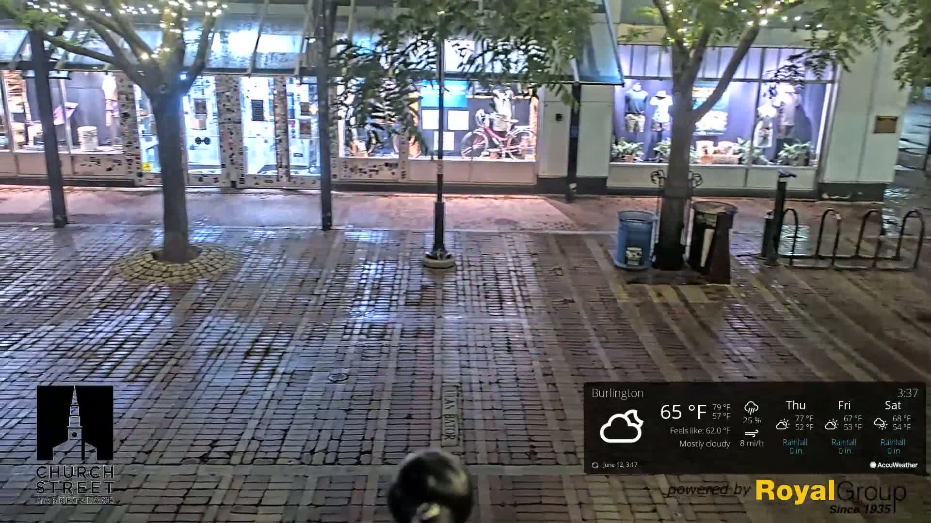 A mostly cloudy night scene shows a brick-paved street in front of storefronts with some trees and bicycle racks, under soft lighting.