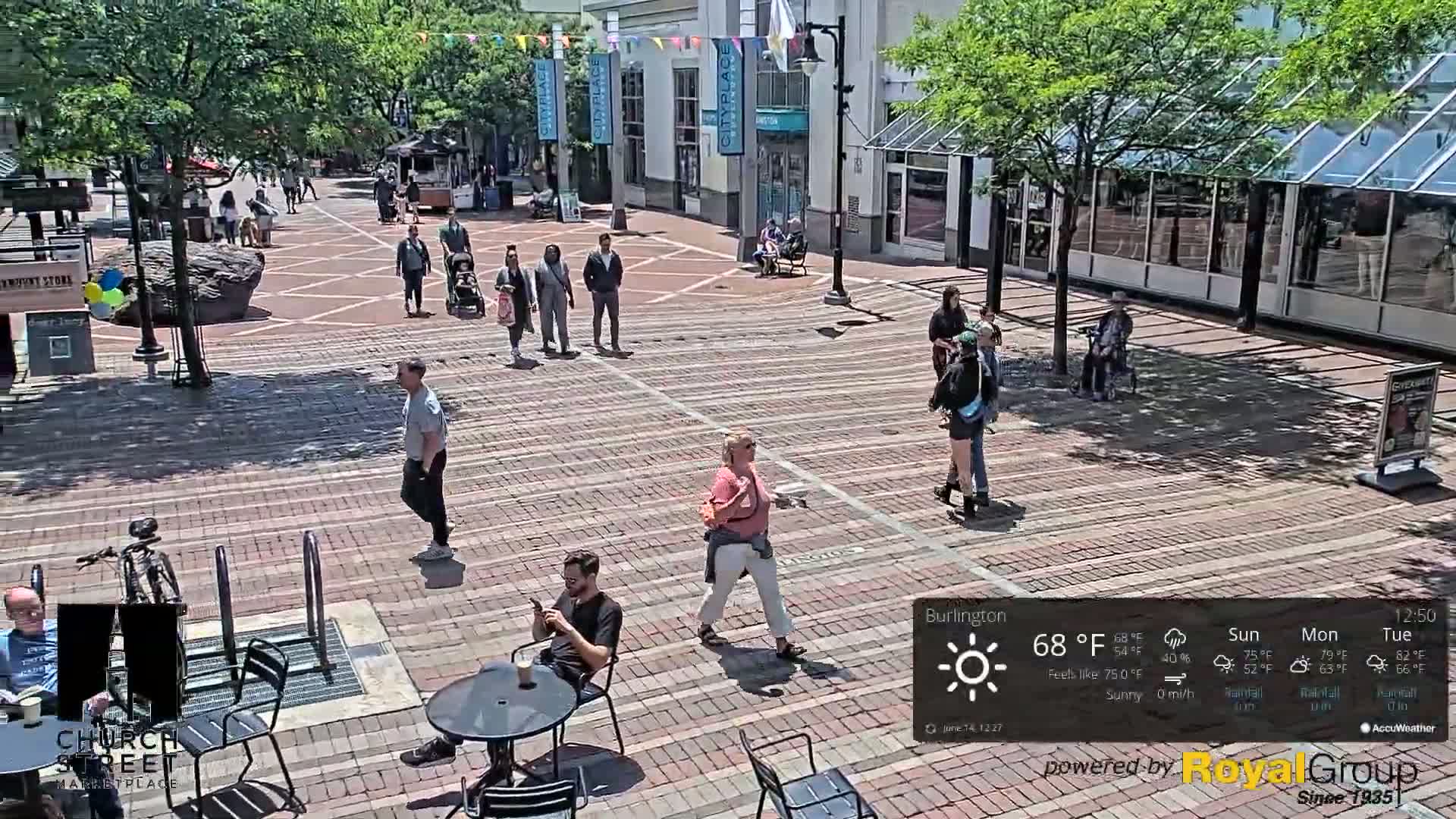 A brick plaza, under sunny skies, features several pedestrians walking among shops and a person sitting at an outdoor table.