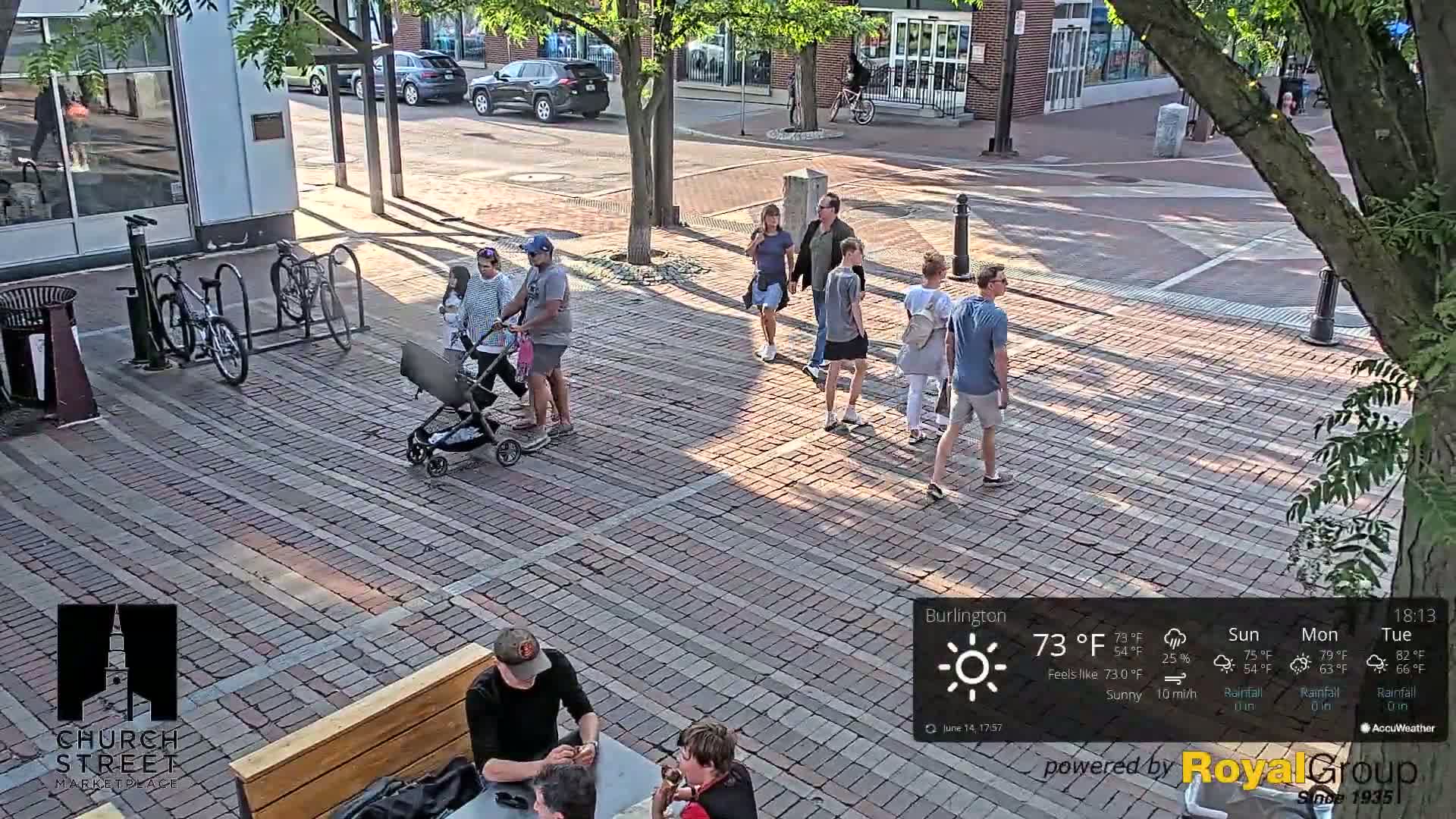 A brick plaza, under sunny skies, features several pedestrians walking among shops and a person sitting at an outdoor table.
