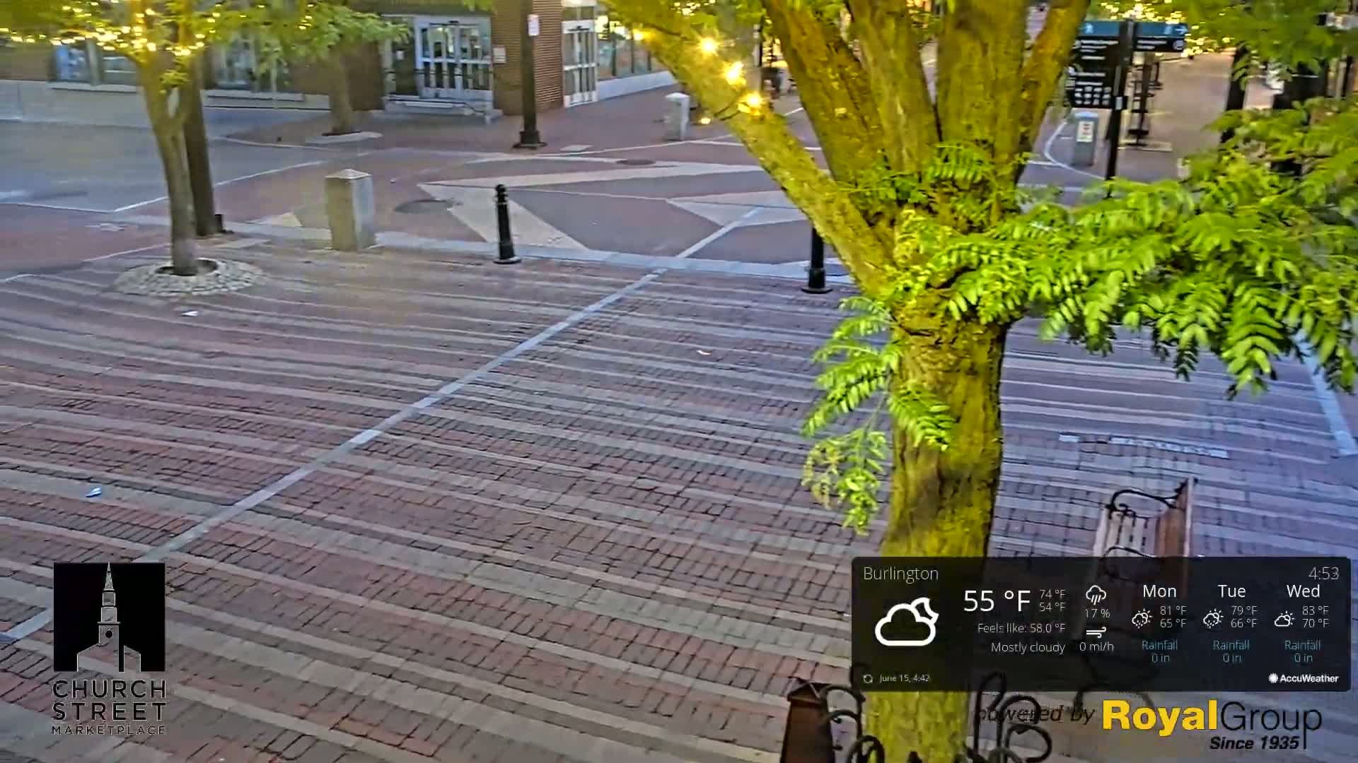 A mostly cloudy evening scene shows a brick paved plaza with a tree in the center, and some buildings visible in the background.