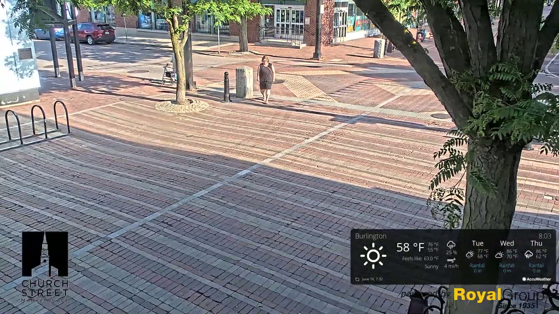 A woman walks across a sunny brick plaza, with a tree and a parked car visible in the background.