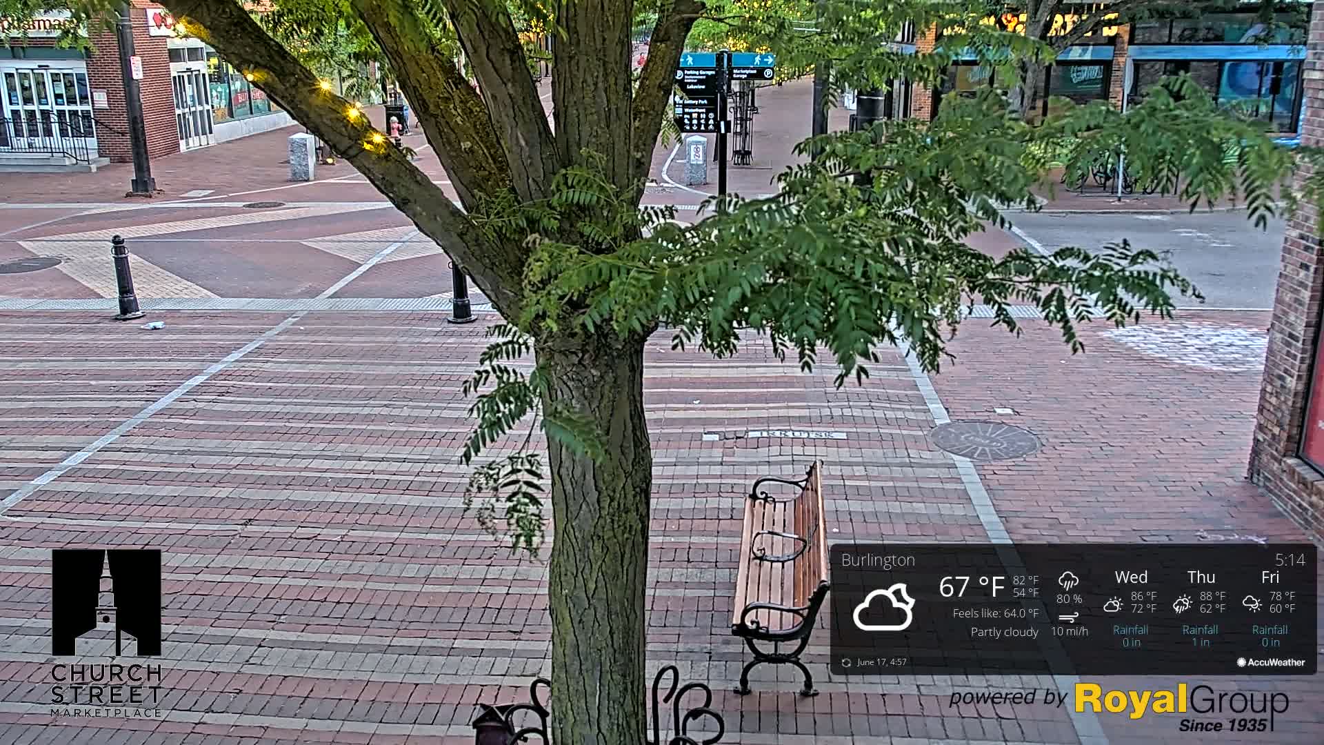 A brick-paved city square with a tree, a park bench, and partly cloudy weather.