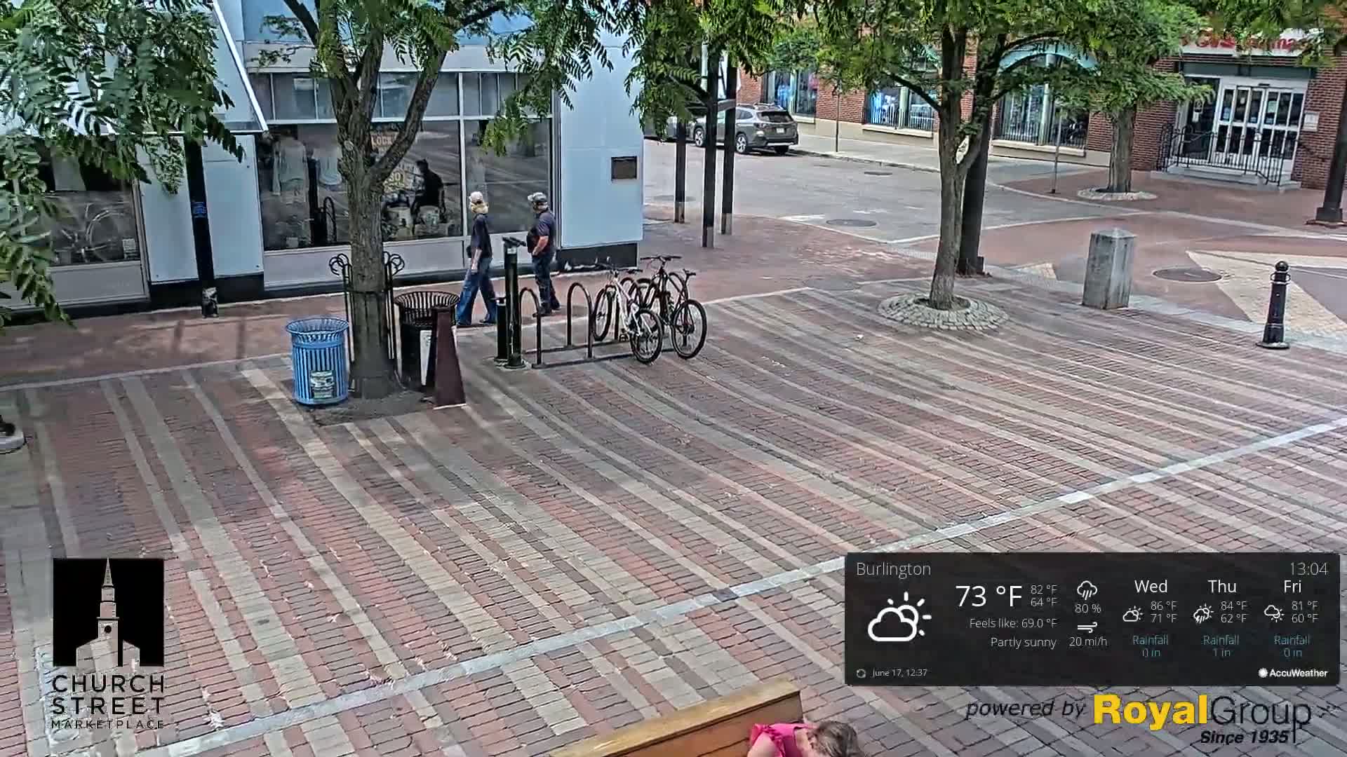 A brick plaza with two bicycles parked at a bike rack, two people walking, and partly sunny weather.