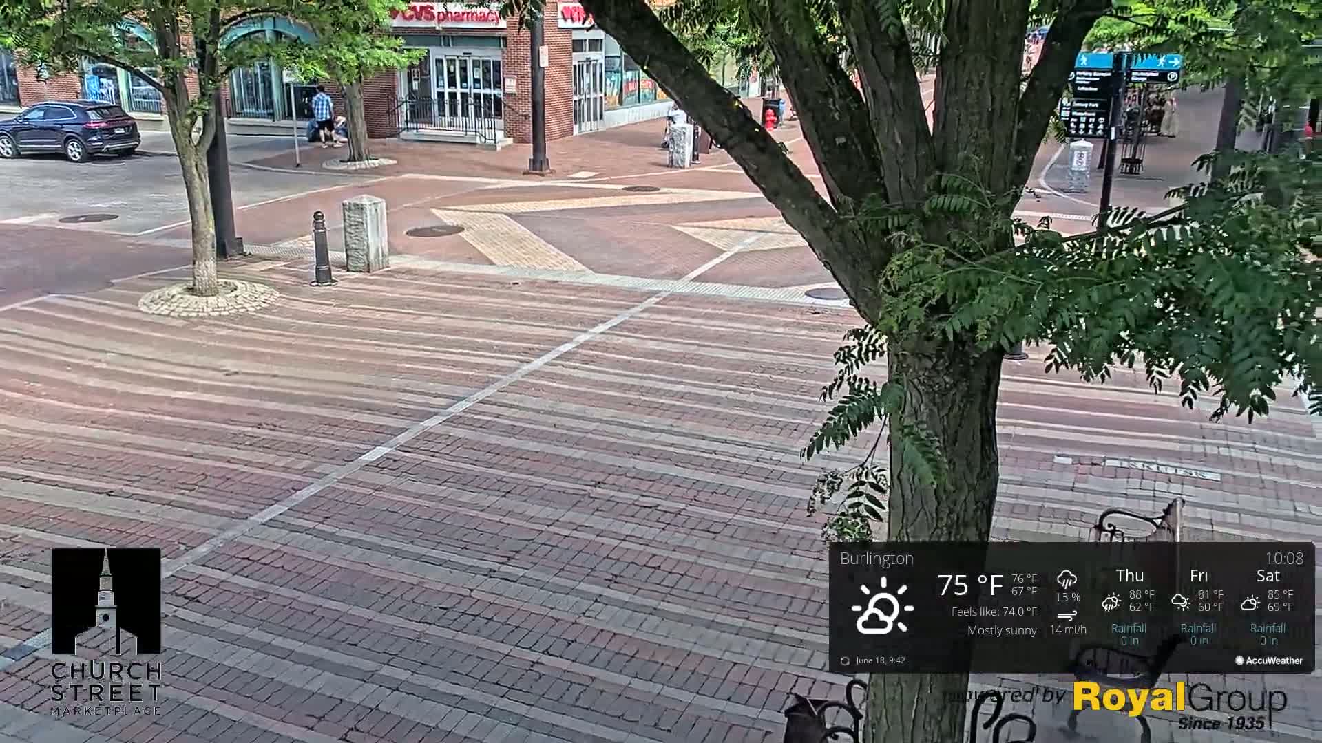 A mostly sunny brick-paved city square is viewed from above, with a large tree partially obscuring the view and a few pedestrians visible in the background.