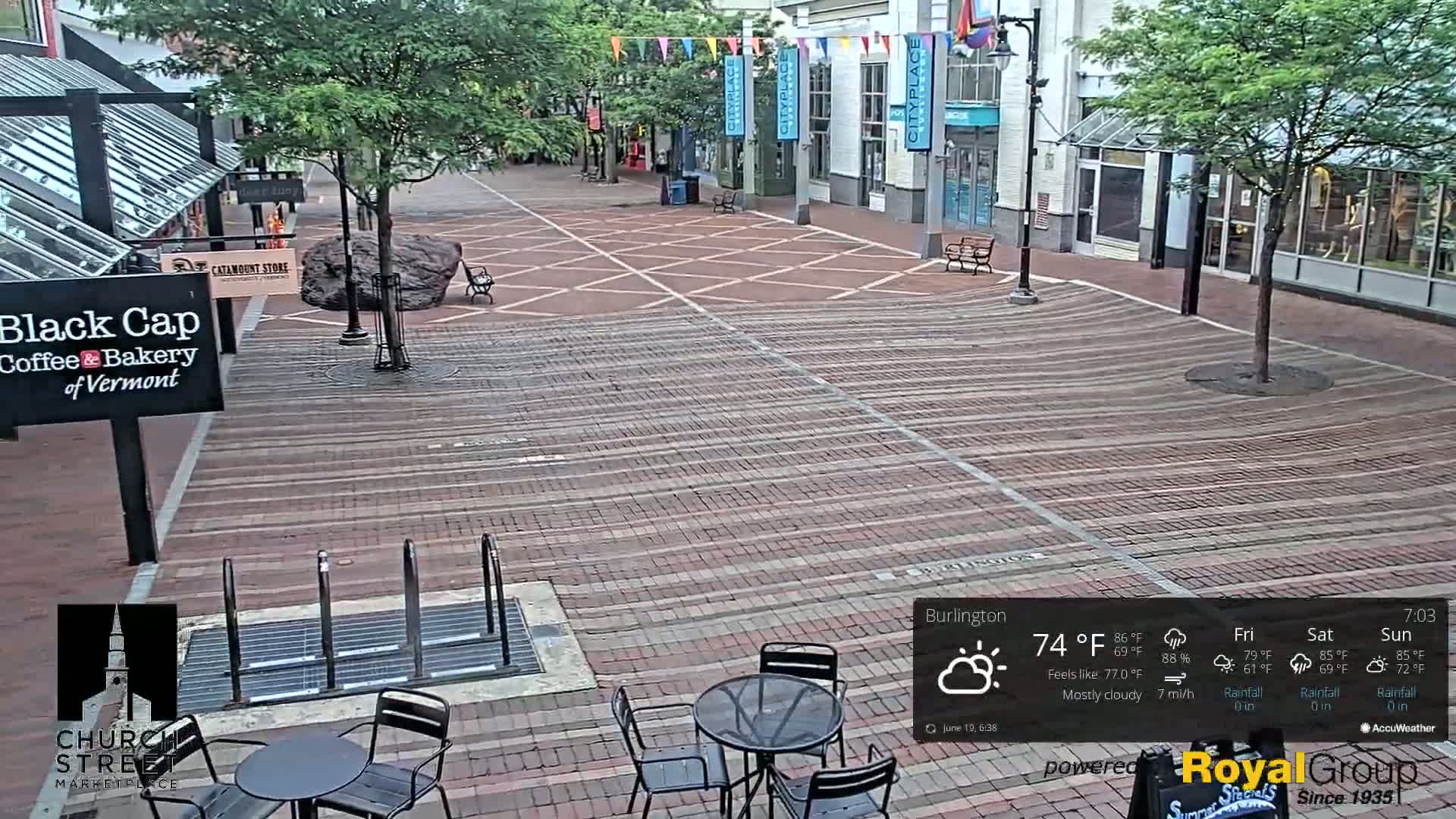 A mostly cloudy outdoor scene shows a brick pedestrian mall with several empty tables and chairs, a large rock, and buildings on either side.