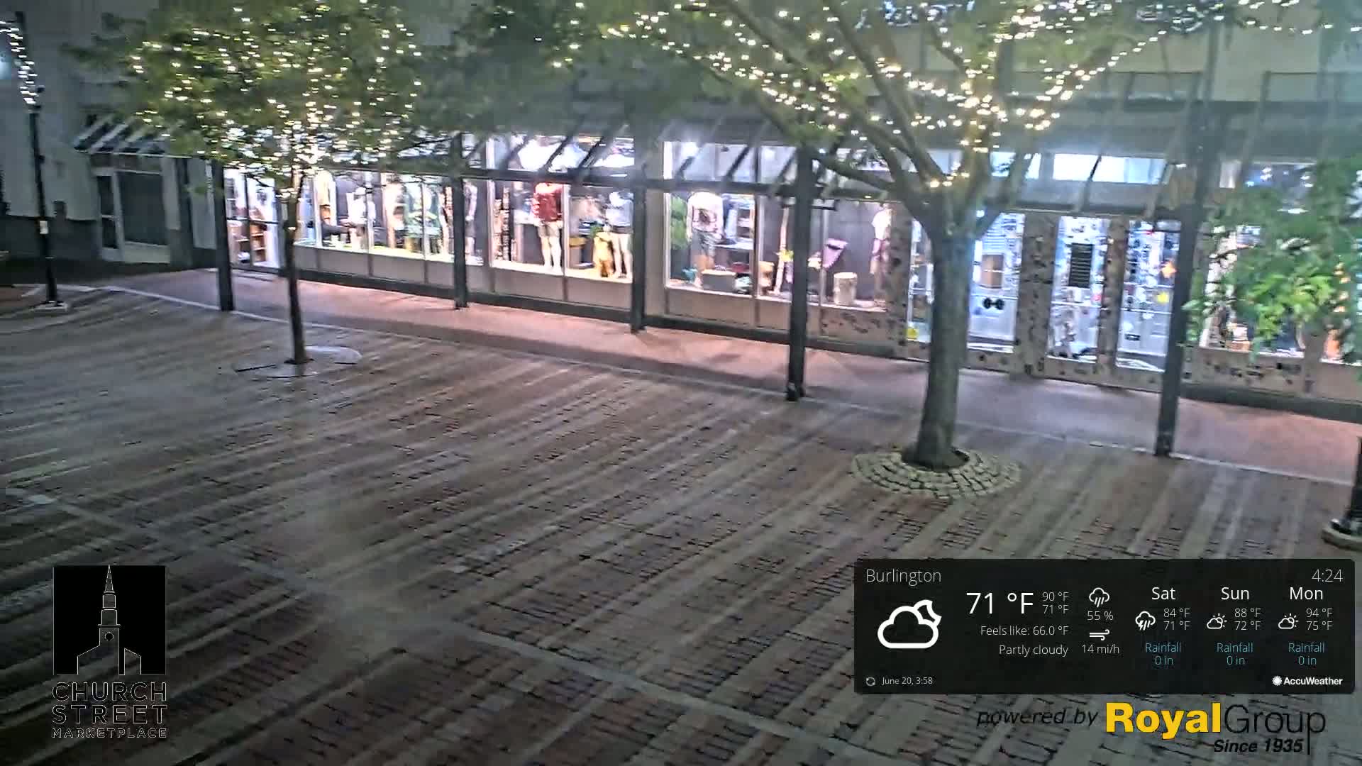 A nighttime view of a brick plaza with a tree strung with lights in front of a store, under partly cloudy skies.
