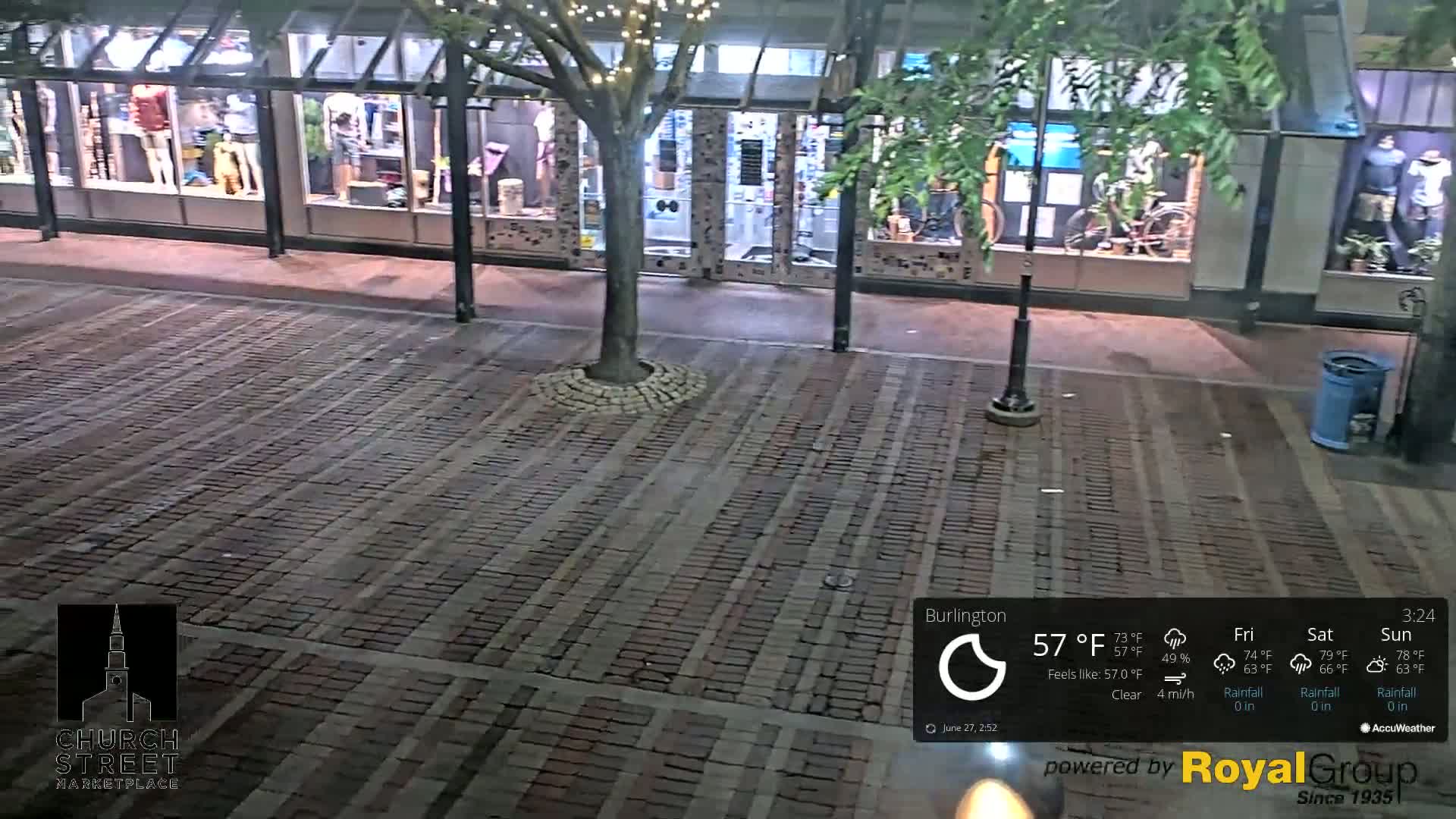 A brick paved area at night is seen, with a tree and storefront windows visible; the weather is clear with a gentle breeze.