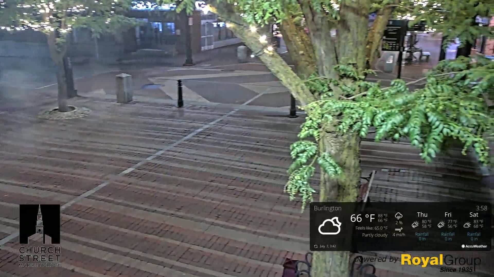 A brick paved plaza at night, partially obscured by a leafy tree, under partly cloudy skies.