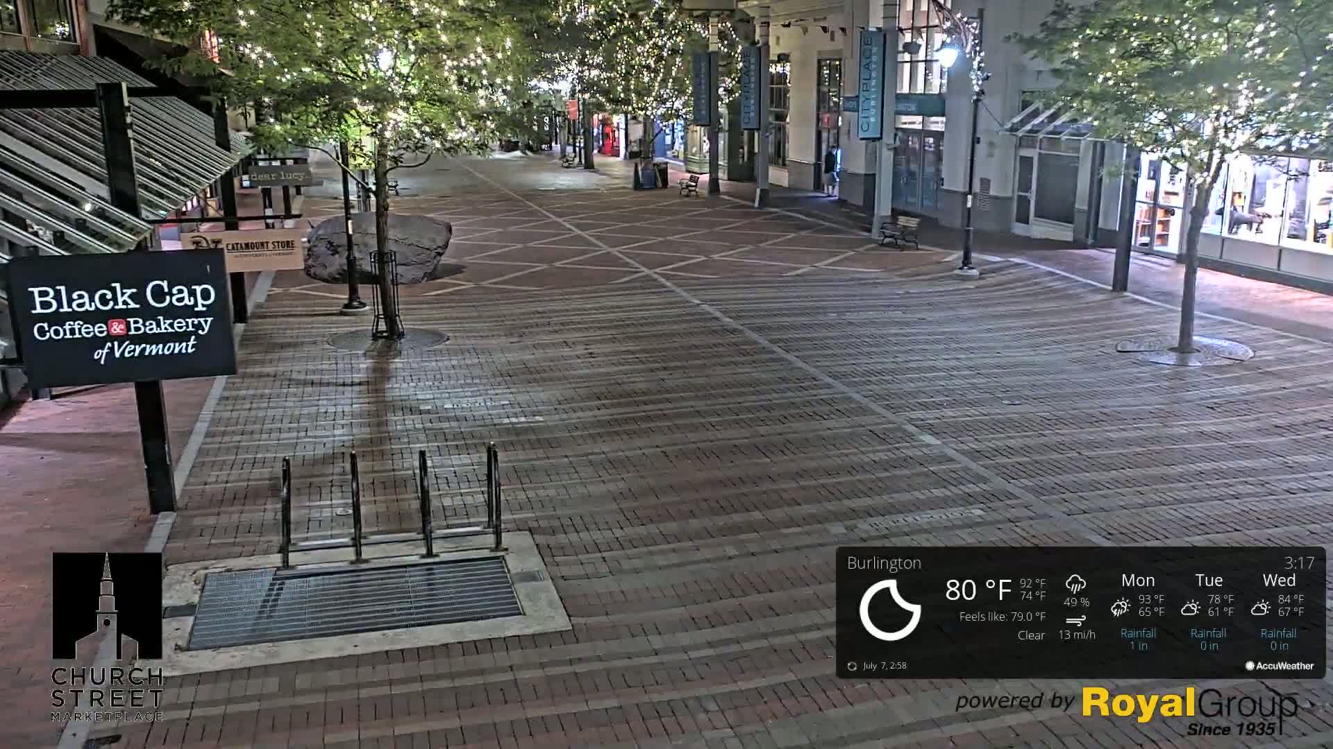 A nighttime view of a brick paved street lined with shops and trees decorated with string lights, under clear skies.