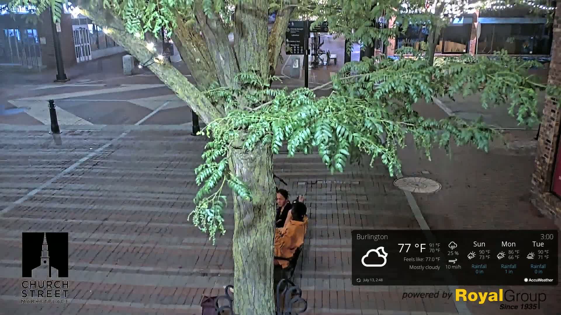 A mostly cloudy night scene shows two people sitting on a bench beneath a tree on a brick-paved street.