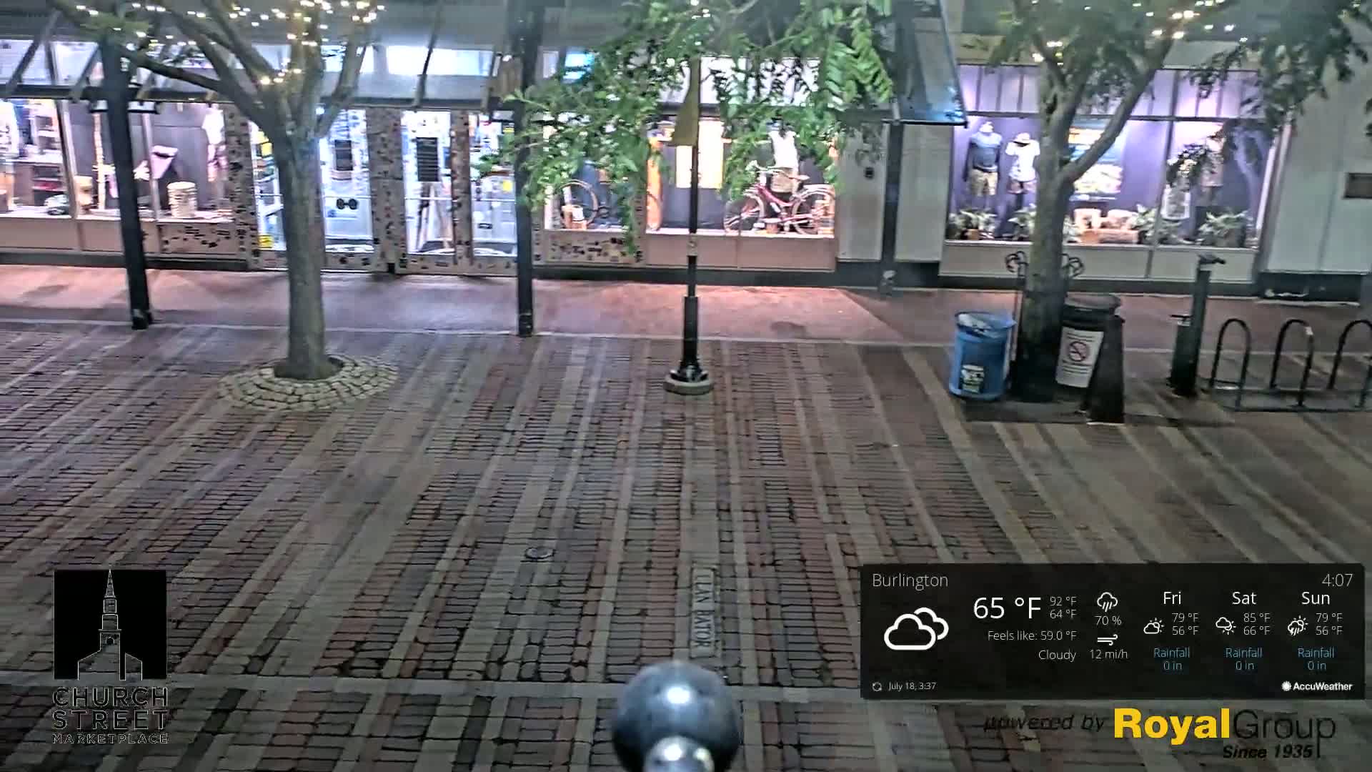 A brick-paved street at night under cloudy skies, lined with shops displaying merchandise in their windows, a tree with string lights, and bike racks.