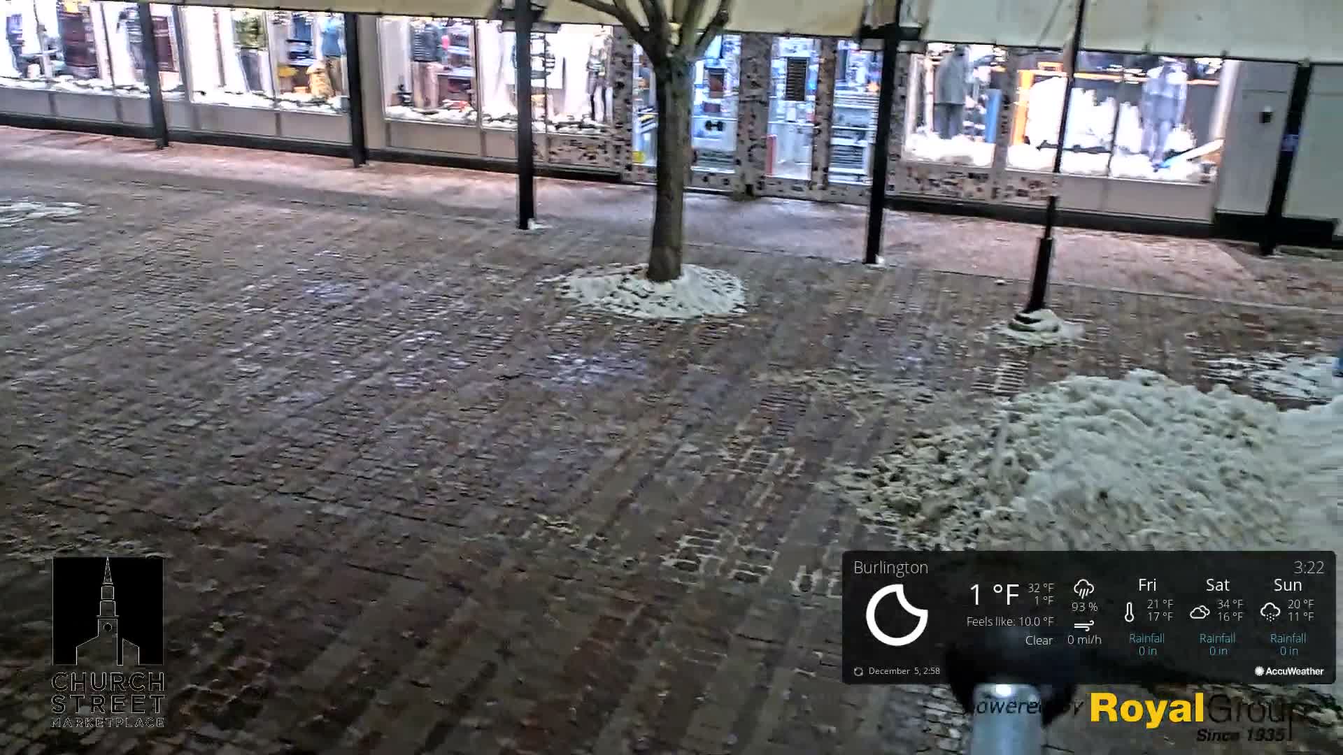 A brick-paved pedestrian street is sparsely covered in snow around a central tree and in larger piles on the right, with illuminated storefronts in the background under clear, very cold 1°F conditions.
