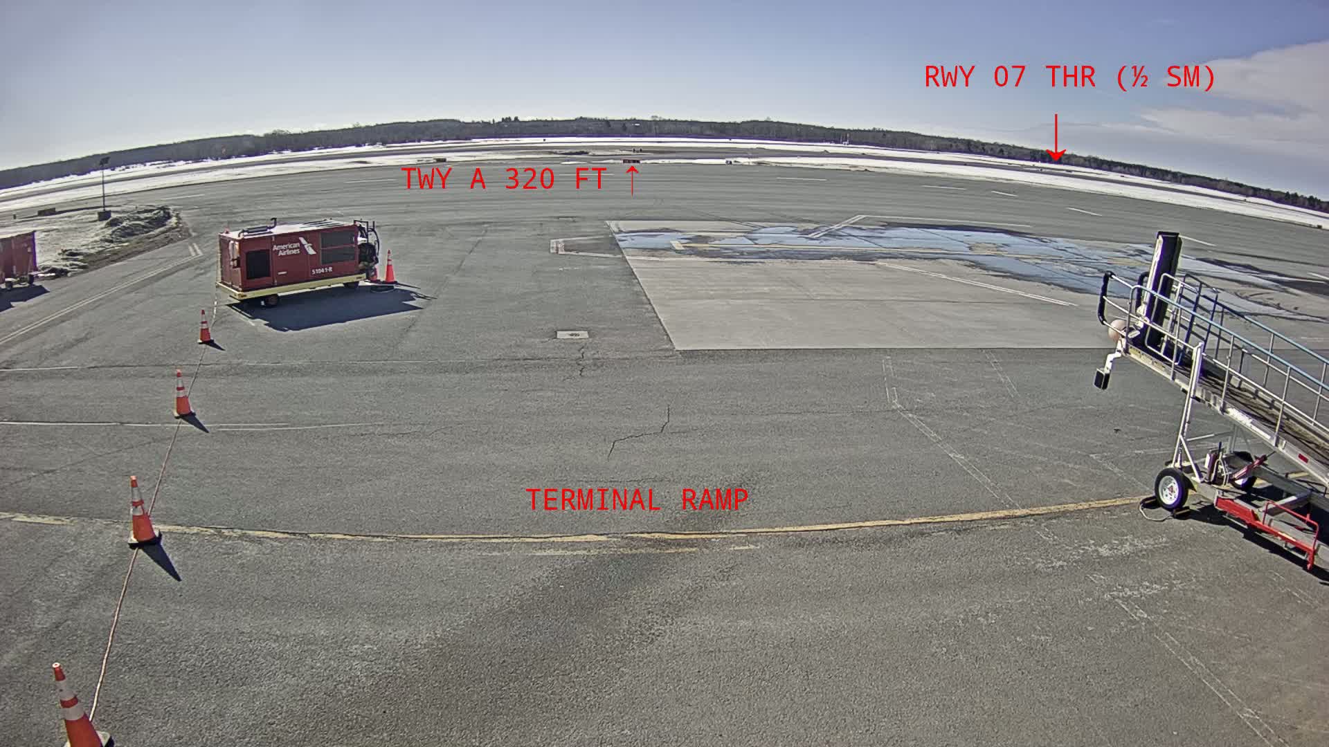 A sunny airport tarmac shows a ground power unit, jet bridge, and several orange traffic cones, with a distant tree line under a clear blue sky.