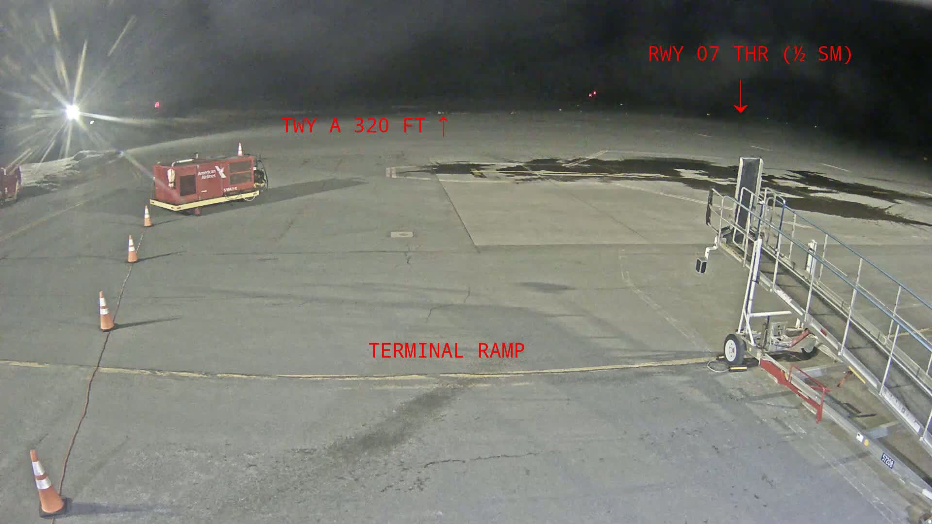 A nighttime airport tarmac scene shows a jet bridge, ground power unit, and orange traffic cones near a terminal ramp.