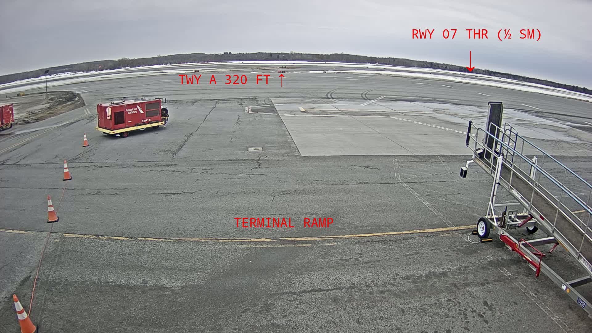 A tarmac with a red baggage tug, a jet bridge, and orange traffic cones is visible on an overcast day.