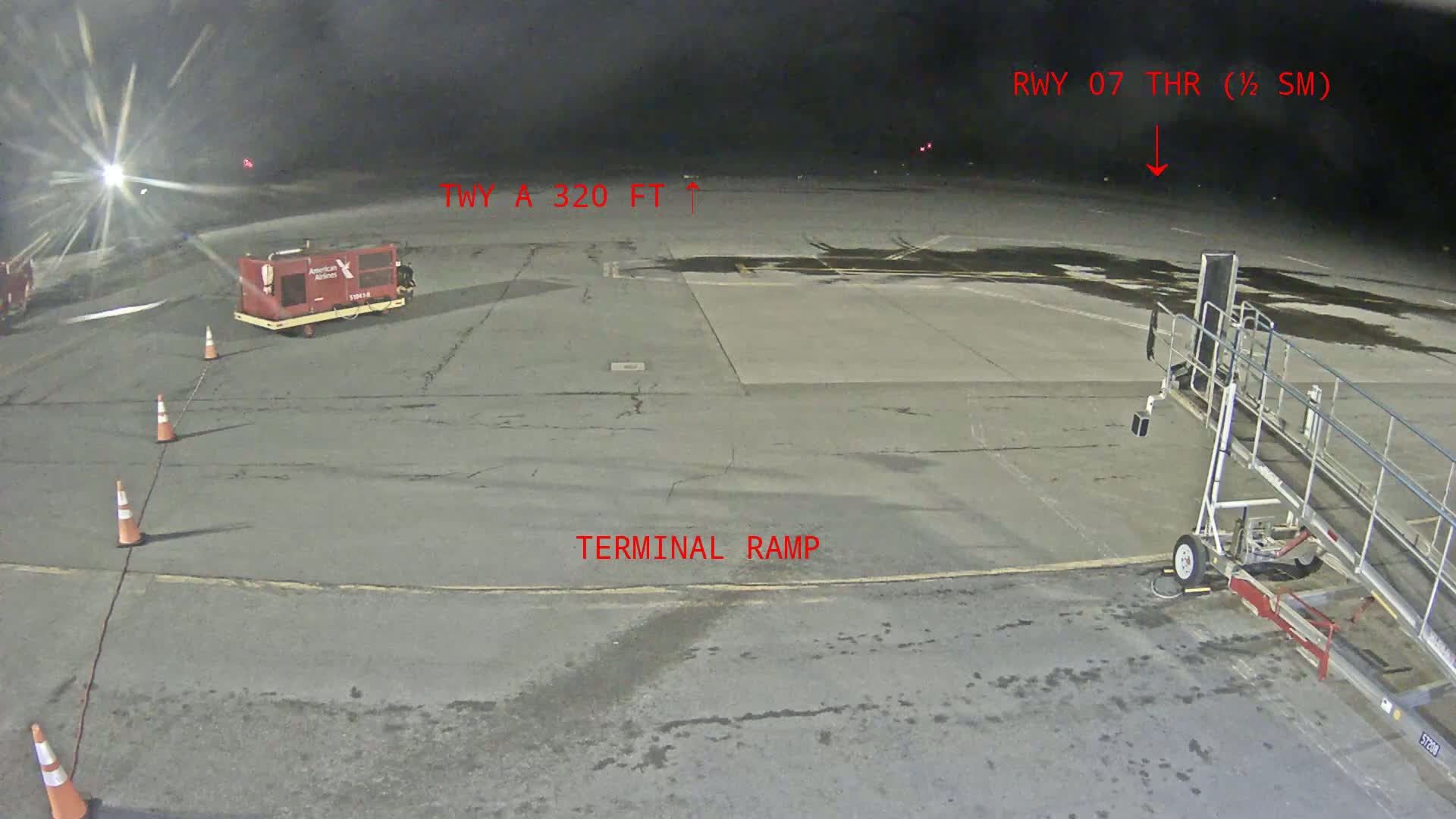 A nighttime view of an airport tarmac shows an empty terminal ramp with a baggage cart and jet bridge, along with several traffic cones.