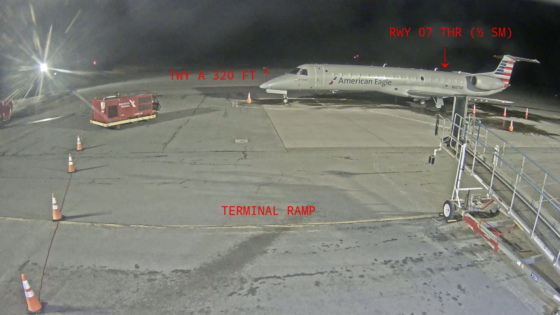 At night, an American Eagle regional jet is parked at a terminal ramp on an airport tarmac, with several orange traffic cones and ground support equipment visible.